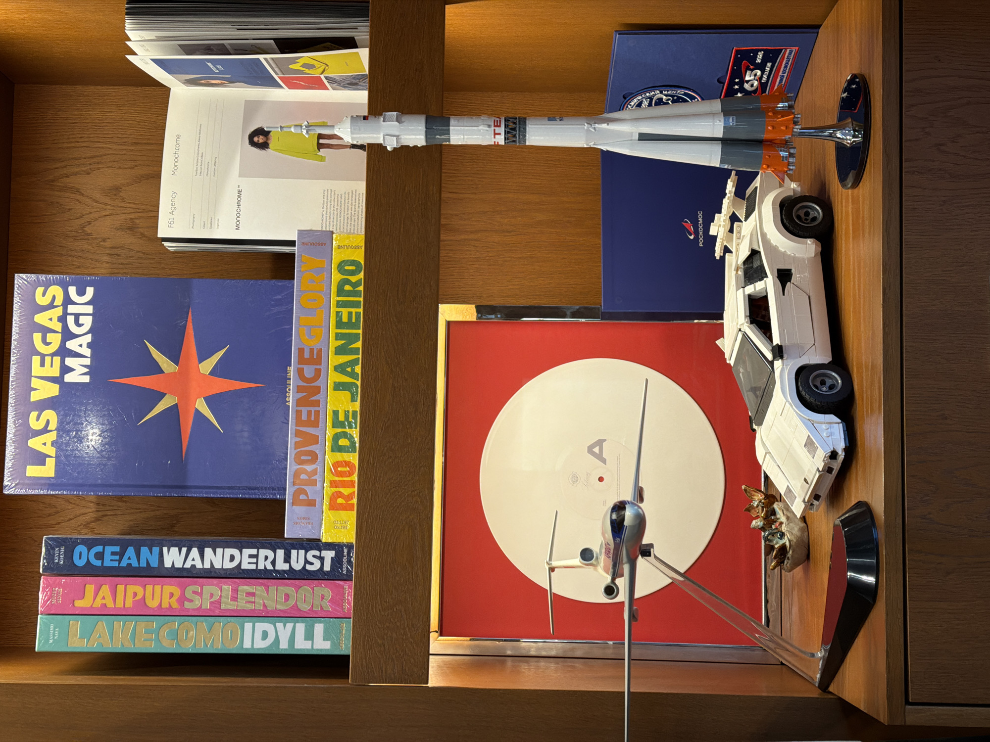 The image captures a wooden bookshelf or display unit in Moscow, Russia, showcasing a curated collection of books and models.
On the top shelf, an open book titled "Monochrome" by "F61 Agency" features a person in a bright yellow garment. Adjacent to it, a detailed model of a Soyuz rocket stands upright, displaying "РОСКОСМОС" (Roscosmos), "ПОЕХАЛИ!" (Poyekhali!), "65", "2026", and "FTE" text. Behind the rocket, a blue folder or box is visible, also bearing the "РОСКОСМОС" logo and "65".
The lower shelf contains a variety of books and collectibles. To the left, a stack of large Assouline travel books includes "LAS VEGAS MAGIC" (with an orange star design), "PROVENCE GLORY," and "RIO DE JANEIRO." Beneath these, three more Assouline books are arranged horizontally: "OCEAN WANDERLUST" by Kevin Koenig, "JAIPUR SPLENDOR" by Noris Singh, and "LAKE COMO IDYLL" by Massimo Mozez.
On the right side of the lower shelf, a white vinyl record with a prominent "A" is displayed in a red frame. In front of the record, a white Lego model of a car, resembling a DeLorean, is placed. Beside the car, a small model airplane is positioned on a transparent stand, and two miniature figures resembling fantastical creatures or gremlins are nestled nearby. The scene depicts a personal arrangement of items suggesting interests in travel, design, space exploration, and pop culture.