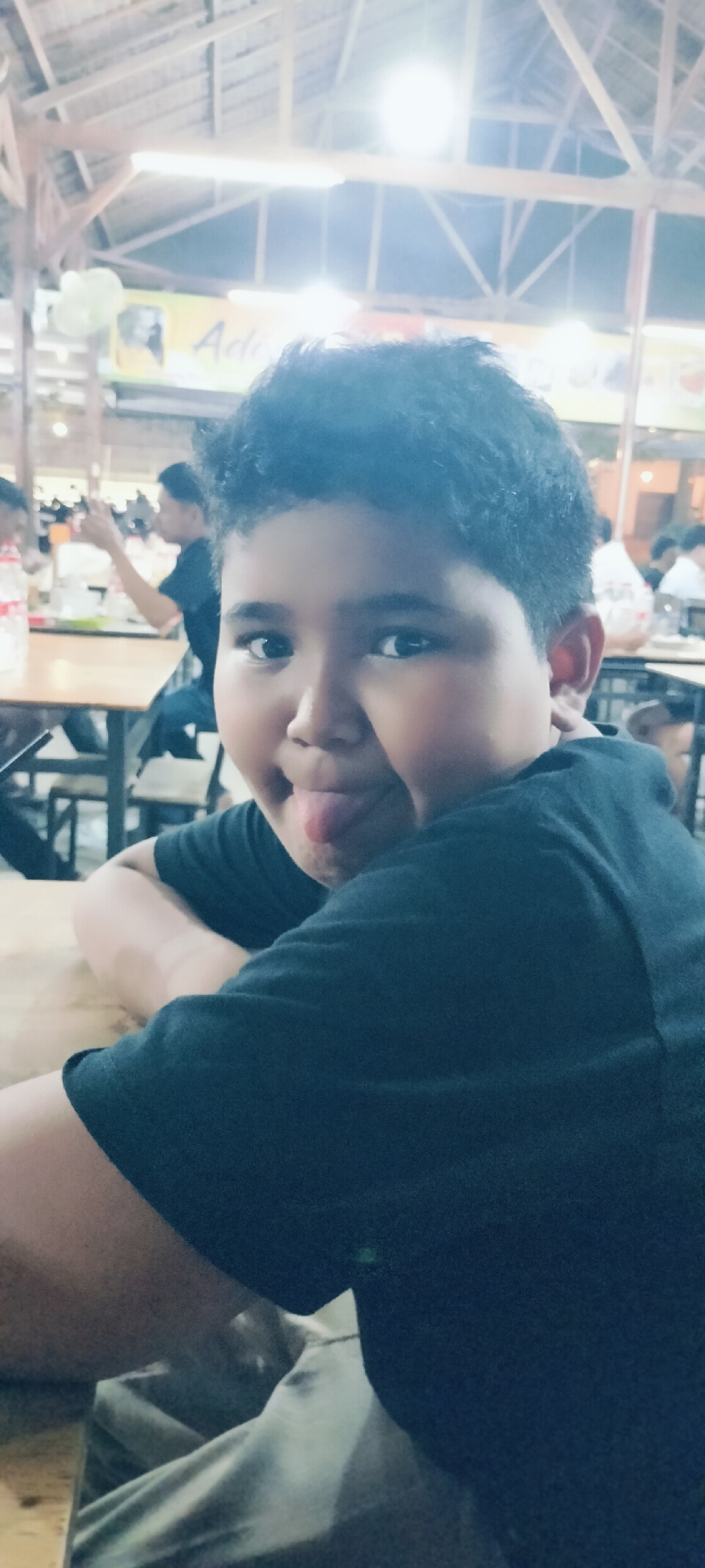 The image features a young boy playfully sticking his tongue out at the camera, seated at a wooden table in what appears to be a casual indoor dining establishment. The boy, who has dark, curly hair and is wearing a dark (likely black) short-sleeved t-shirt, is the central focus, resting his arms on the table. His expression conveys a sense of lightheartedness or mischief.
The setting suggests a restaurant, cafe, or food stall, consistent with the location context of Banda Aceh, Indonesia. In the blurred background, several other people are visible seated at wooden tables, indicating a social or dining environment. A partially visible plastic bottle with a red label sits on the table to the left. The roof structure above is characterized by exposed wooden beams and trusses, giving the space a rustic or traditional feel. Bright artificial lights are prominent overhead, causing some glare and lens flare, which implies the photo was taken in the evening or at night. A white ceiling fan is partially visible in the upper left background.
A yellow banner can be seen in the upper left, behind the main subject, with the word "Ade" clearly legible in a stylized font. This could be part of the establishment's name or an advertisement. The overall scene depicts a relaxed moment in a local eatery.