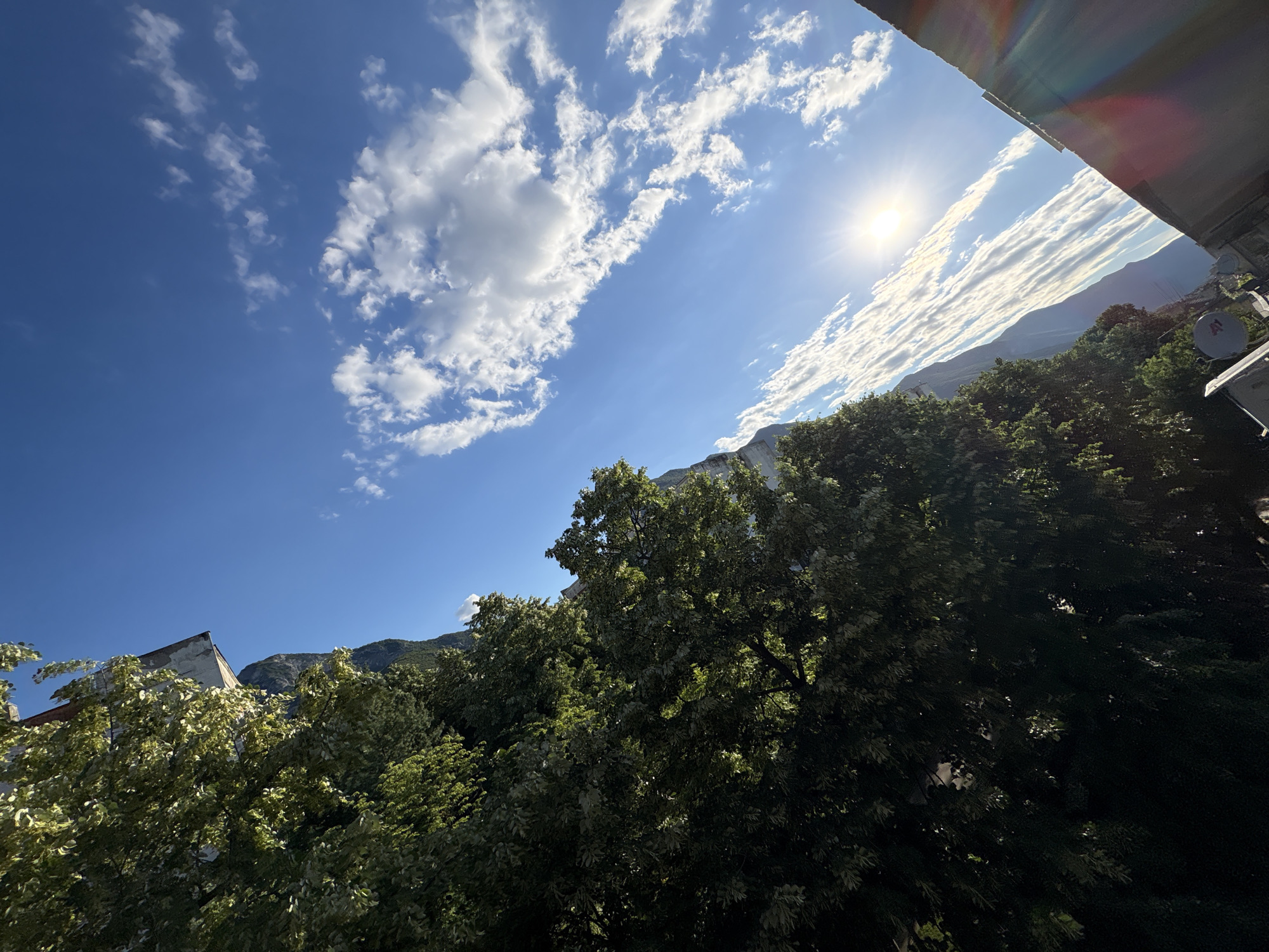 The image depicts a bright, sunny day in Vratsa, Bulgaria, with a view angled towards the sky and mountains. Lush green trees dominate the left side of the frame, their leaves illuminated by the sun. In the upper left corner, a portion of a building with a faded facade is visible, partially obscured by the foliage. Beyond the trees and building, a majestic mountain range stretches across the background, its peaks shrouded in shadow and mist. The sky is a vibrant blue, dotted with fluffy white clouds that are elongated and streaky, suggesting wind activity. The sun is a radiant orb, positioned in the upper right quadrant of the image, casting a warm glow and creating lens flares that streak across the lower right corner, suggesting the photograph was taken with a digital camera. A portion of a rooftop or balcony railing, casting a shadow, is also visible in the lower right corner. The overall scene conveys a sense of natural beauty and tranquility, with the urban elements subtly integrated into the landscape.