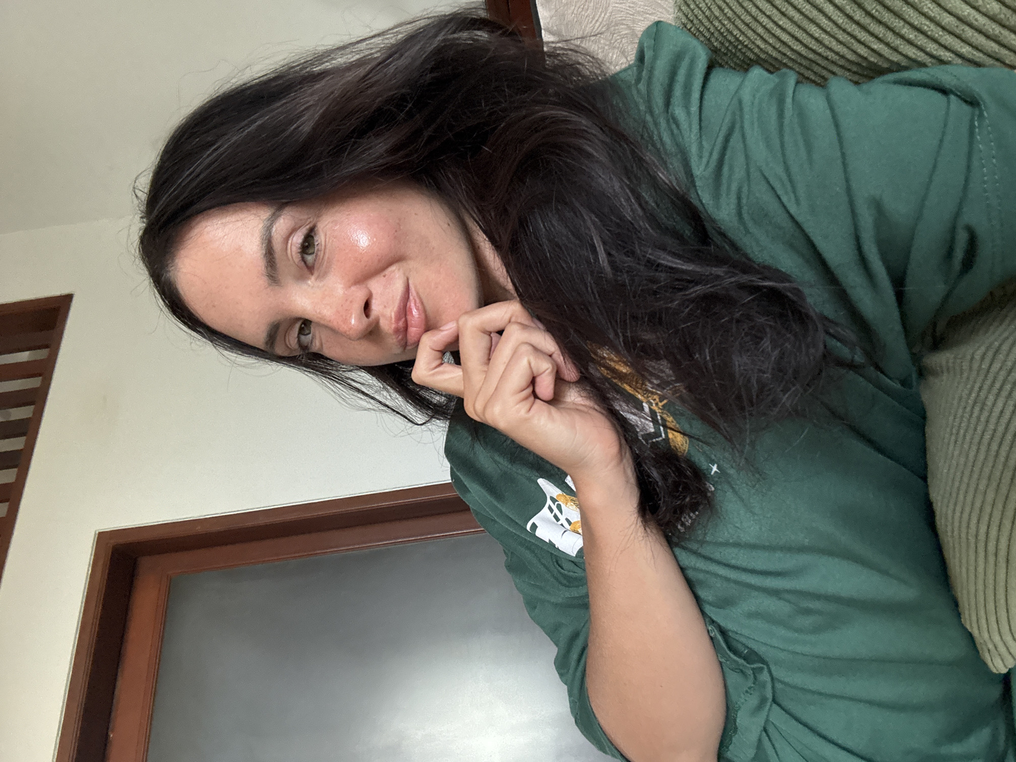 A woman with dark hair and fair skin is captured in a selfie, resting her chin on her hand and looking directly at the camera. She is wearing a dark green t-shirt and is positioned on a couch with a ribbed texture. Behind her, a wooden-framed door with a frosted glass panel is visible, along with a section of a white wall and a wooden ceiling trim. The scene appears to be indoors, potentially in a residential setting in North Kuta, Indonesia.