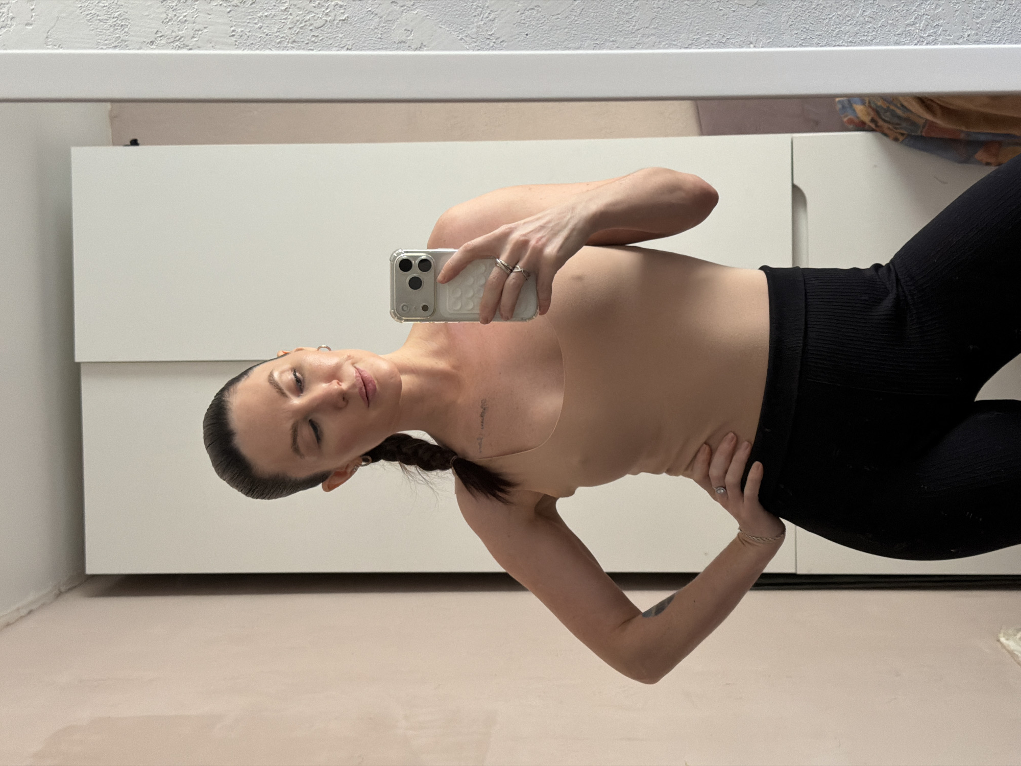 A woman is taking a selfie in what appears to be an indoor setting. She is wearing a tan-colored top and black leggings. The background includes a light-colored wall with a cabinet or drawers, and a light-colored floor. There is no information to suggest the photo was taken in the city of Perm, Russia.
