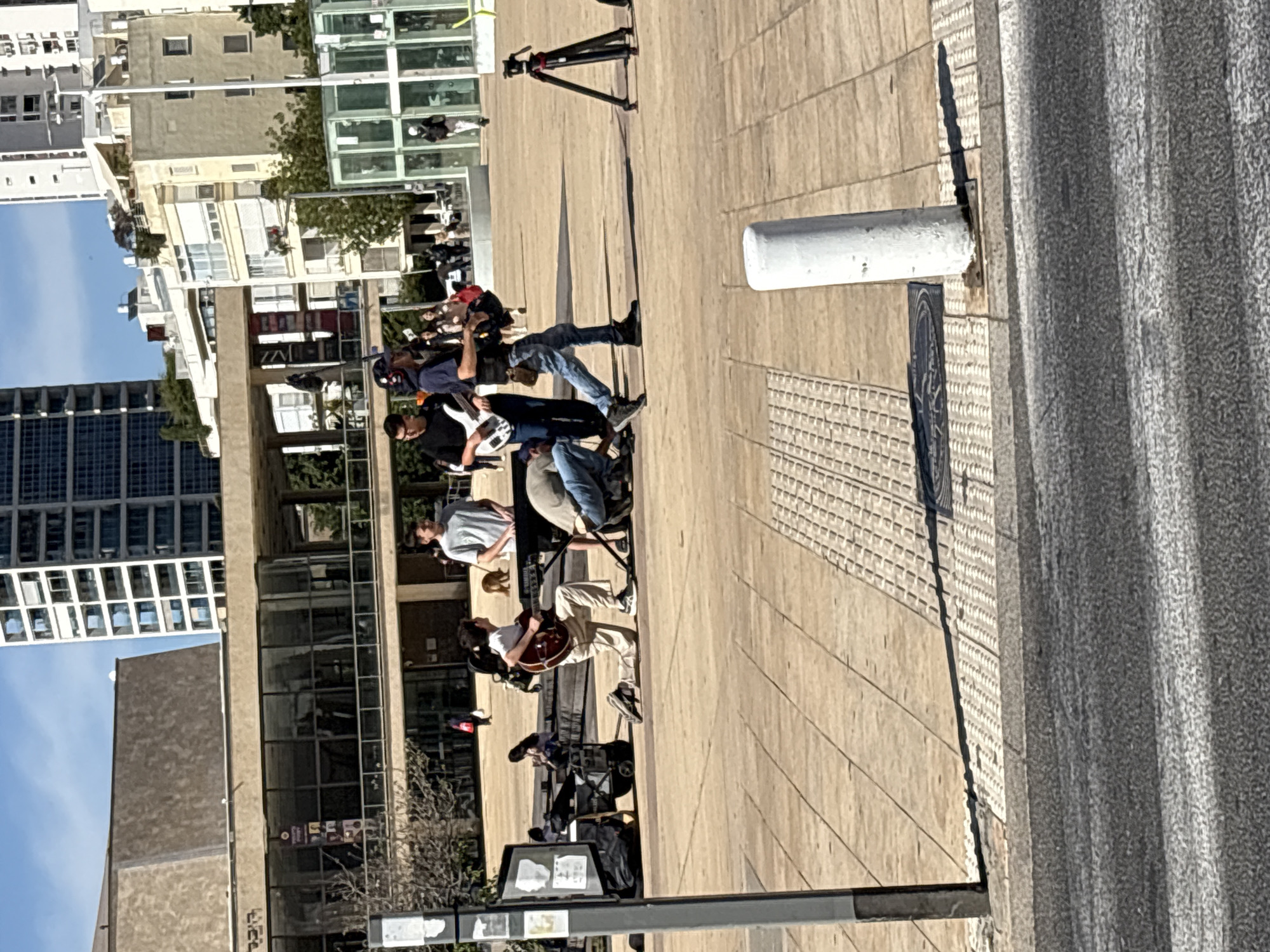 A group of street musicians performs on a paved area in Tel Aviv, Israel, as evidenced by the architecture and street furniture visible. The group consists of at least four individuals, each playing an instrument, including what appears to be guitars and possibly a bass. They are set up with musical equipment, including amplifiers and stands. A camera on a tripod is positioned nearby, possibly for recording their performance. Buildings are visible in the background, including a taller modern structure with reflective windows. The scene is bathed in daylight, casting shadows across the paved surface.