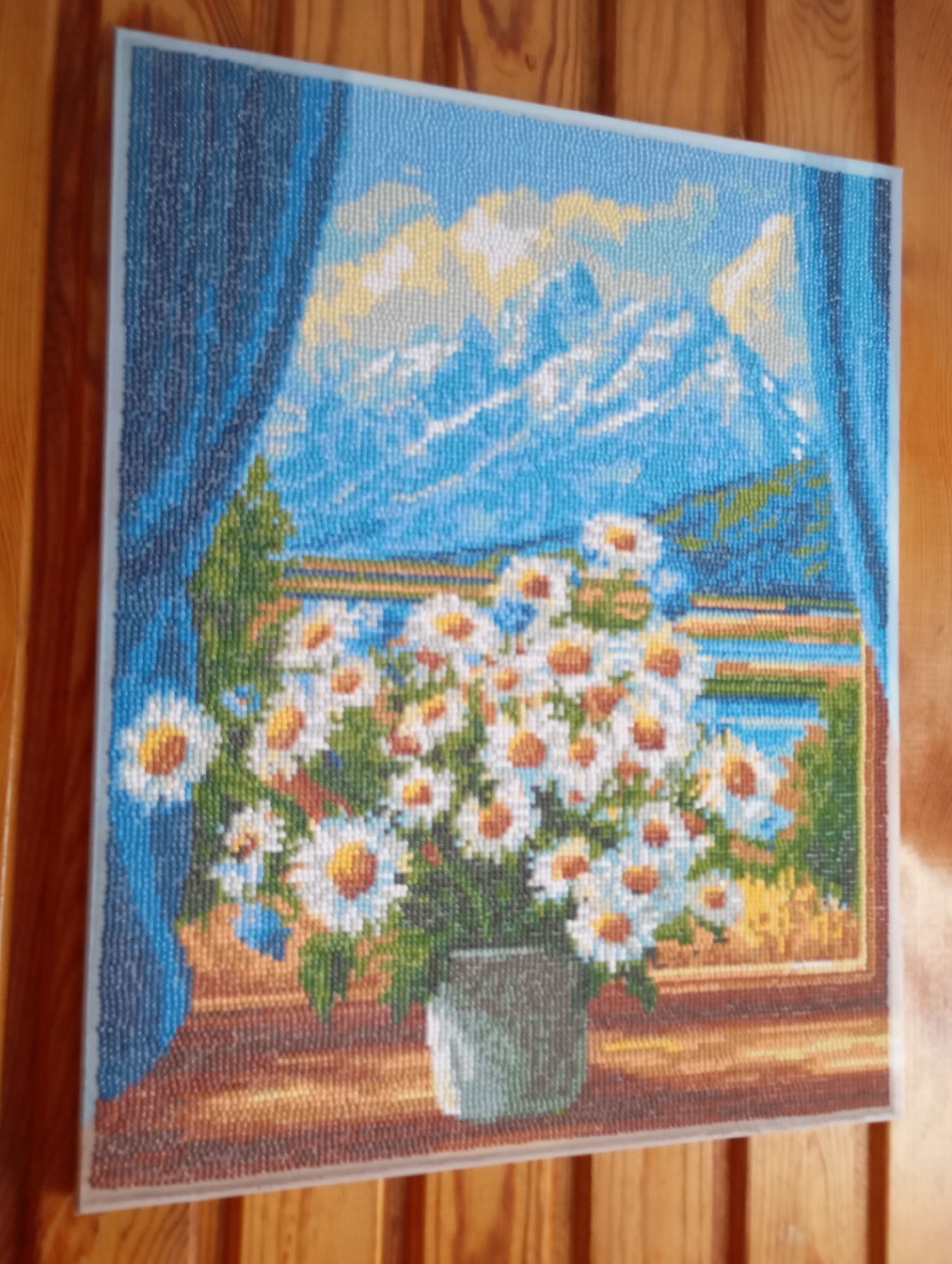 A framed diamond painting depicting a still life of daisies in a vase on a windowsill, with a mountain landscape visible through the window, hangs on a wooden wall. The painting features a bouquet of white daisies with yellow centers in a gray vase. Behind the vase, a body of water is visible with mountains in the distance, under a sky with yellow and white clouds. A blue curtain is draped on the left side of the painting, framing the scene. The diamond painting is oriented vertically and is set against a backdrop of vertical wooden planks. The location specified, Льниха, Russia, is not discernible from the image itself.