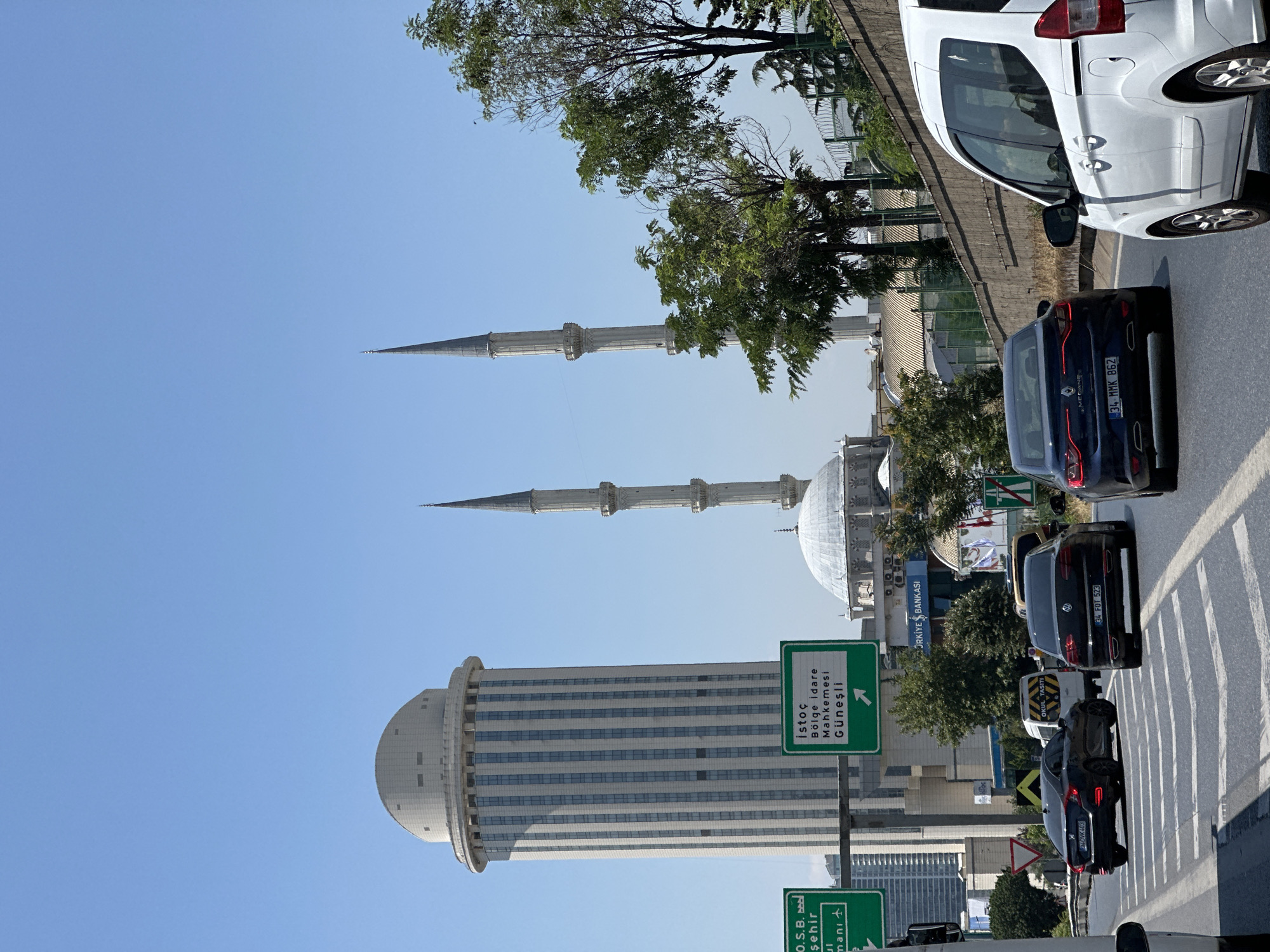The image shows a street scene in the city of Bağcılar, Turkey, on a clear day. In the background, a large, modern, cylindrical building with a domed top stands tall. Adjacent to it, a mosque with a dome and two minarets is visible. Several cars are on the road, moving in the same direction, indicating traffic. Some of the cars are black sedans, and a white van is also present. Green road signs with white lettering are placed on poles, providing directions. Trees line the side of the road, and a concrete wall is also present. The sky is bright blue. The overall scene depicts an urban environment with a mix of modern architecture and religious structures.