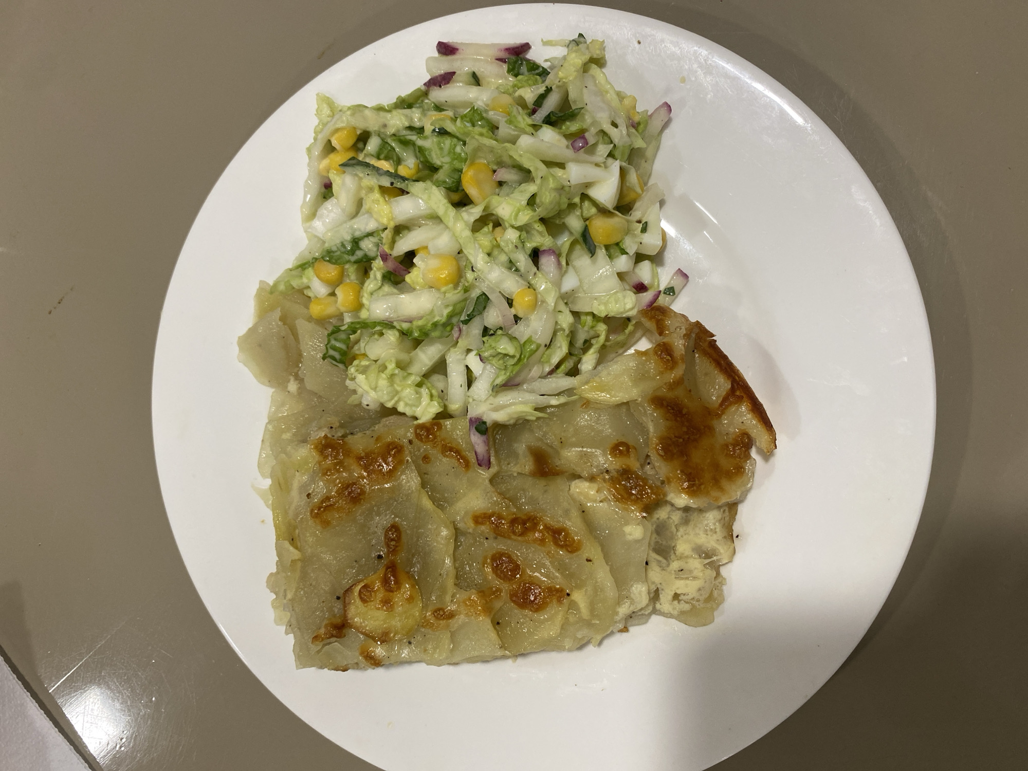 The image shows a white plate with a meal on it, placed on a light brown surface. The meal consists of two distinct parts. On the left is a slice of what appears to be a gratin or casserole, made of thinly sliced potatoes layered and baked with cheese on top, giving it a golden-brown, slightly crispy appearance. The potatoes are tender and some appear translucent, with melted cheese clinging to them. Some black specks, likely pepper, are visible on the potato layers. The right side of the plate features a fresh salad. This salad is composed of shredded lettuce or cabbage, along with pieces of corn kernels, chopped red onion, and possibly some diced hard-boiled egg or other white ingredient. The salad is dressed in a creamy white dressing.
The meal is positioned centrally on the plate, with ample white space around it. The lighting in the image is direct and somewhat harsh, casting subtle shadows. The background is a neutral, uniform light brown. The surface on which the plate rests appears to be a table or countertop with a slightly textured, matte finish. There are no visible people or any indication of activity in the image, making it a still-life composition focused solely on the food. There is no information in the image to suggest its location in Rozhny, Ukraine.