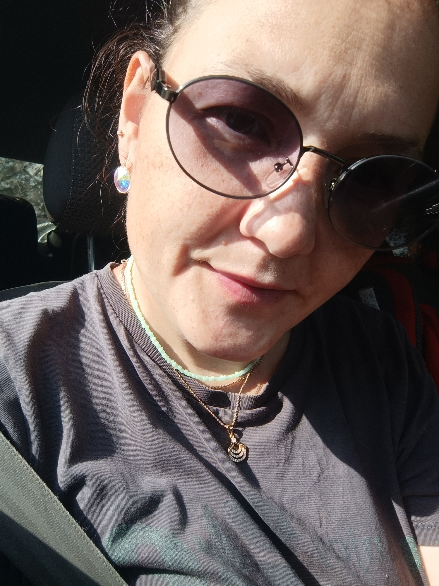 A woman with light brown hair, wearing sunglasses and a grey t-shirt, is seen from the chest up, inside a vehicle. She has a light skin tone and is wearing a light green beaded necklace and a gold pendant necklace. She also has a dangling earring in her left ear. The sunlight is casting shadows on her face and t-shirt. The image is a close-up and does not show the surrounding environment in detail, so it is impossible to determine her location in the city of Foki, Russia.
