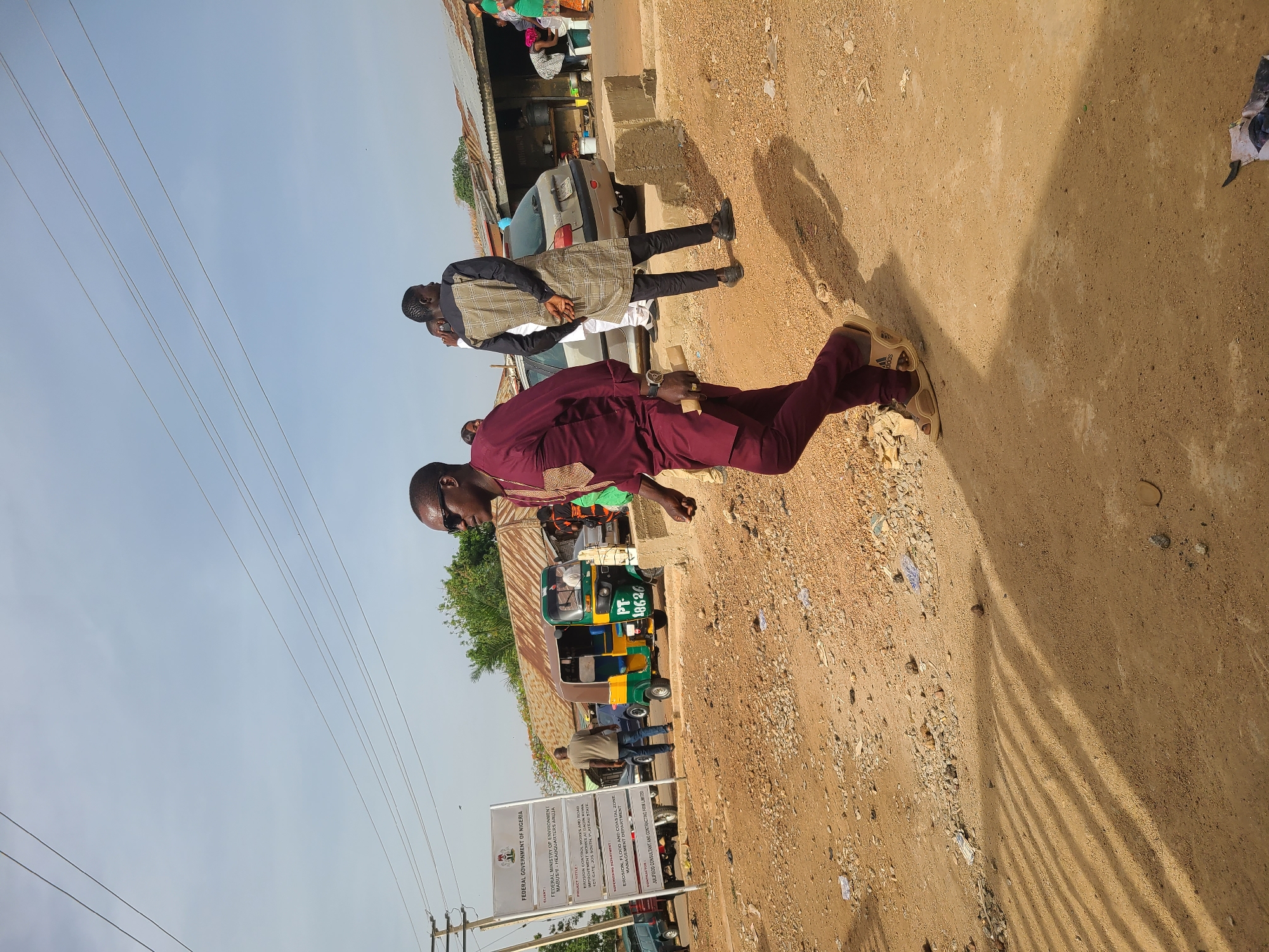 The image shows a dusty street scene, likely in a residential area, on a sunny day in Jos, Nigeria. A man in a maroon suit and sunglasses is walking down the street. Another man dressed in a white shirt and a dark plaid outer vest is seen nearby a light-colored car. A green and yellow tricycle is parked near a sign indicating "FEDERAL GOVERNMENT OF NIGERIA, FEDERAL MINISTRY OF ENVIRONMENT, MABUSHI HEADQUARTERS ABUJA." The sign also notes the project title "EROSION CONTROL WORKS AND ROAD IMPROVEMENT WORKS AT DA TET CATE, JOS SOUTH, PLATEAU STATE". Powerlines run overhead. The ground is dirt and gravel, with some scattered debris.