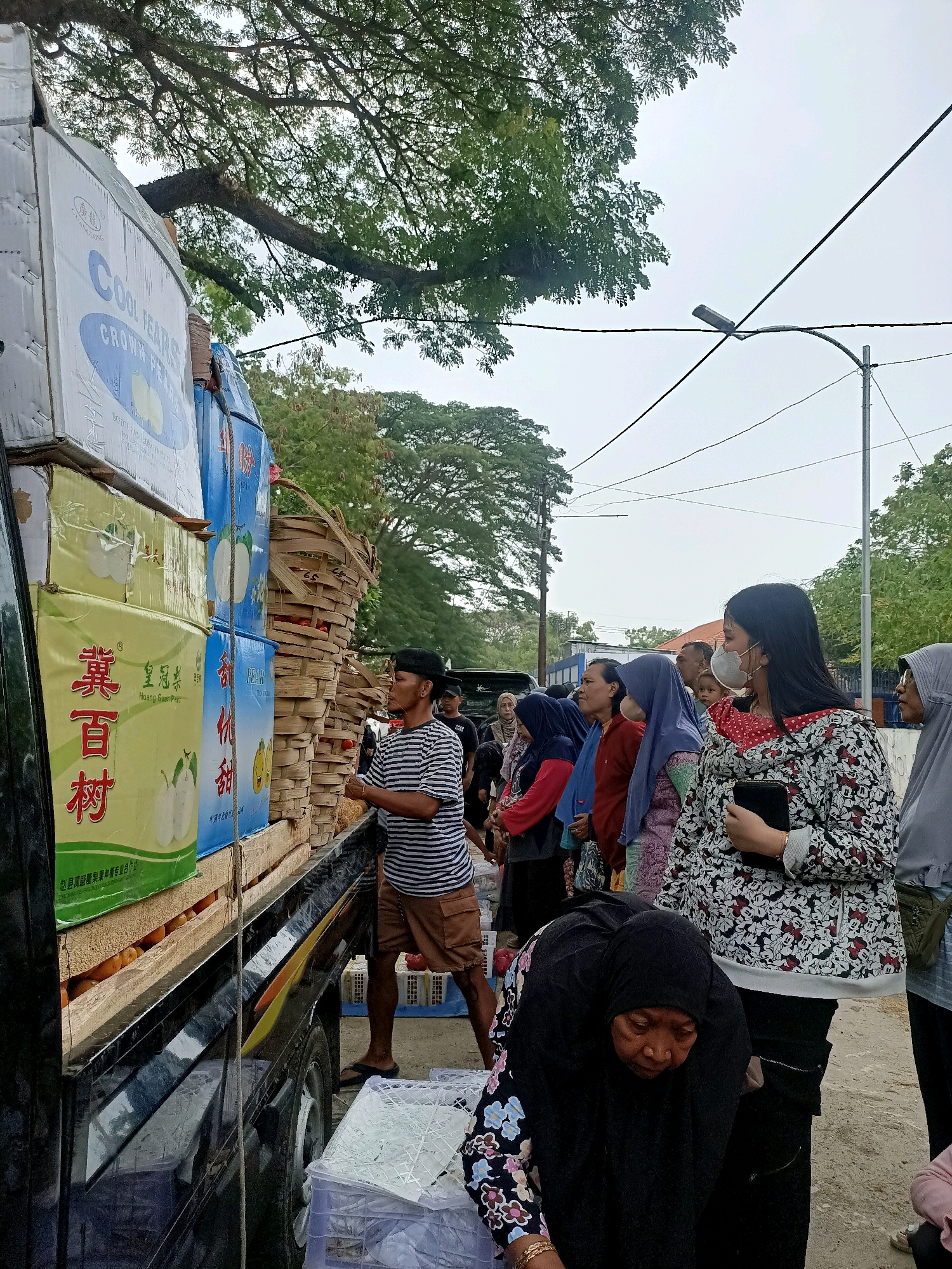 In Madiun, Indonesia, a scene unfolds as a group of people are gathered around a truck laden with boxes of fruit. The boxes are stacked high on the back of the truck, some bearing labels in Asian script. In the foreground, a man wearing a striped shirt and shorts is handing something from the truck. Several women, some wearing hijabs, are standing in a line, appearing to be waiting for their turn to receive the goods. One woman in a floral patterned shirt and a face mask is holding a black object, possibly a phone or wallet. Another woman in a black hijab is bent over, looking down. The environment appears to be outdoors, with trees and power lines visible in the background. The ground is a dusty, sandy surface. The overall atmosphere suggests a distribution of goods, possibly a market or a community event.