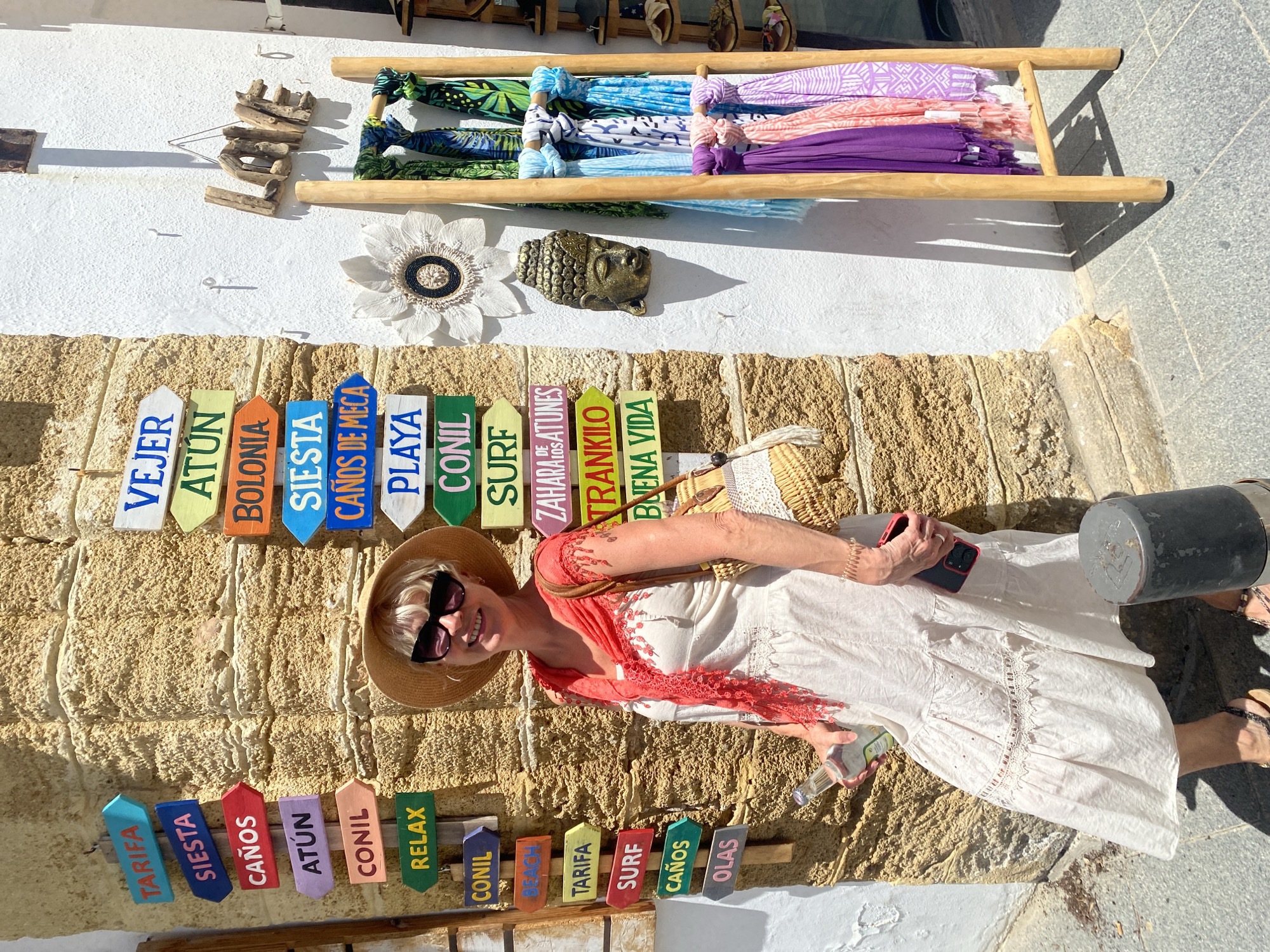 In Conil de la Frontera, Spain, a woman with blonde hair and sunglasses, wearing a white dress and a straw hat, stands against a textured stone wall. She holds a bottle in her left hand and a smartphone in her right. A woven straw bag hangs on her shoulder. The wall is adorned with colorful directional signs pointing to various locations including "Tarifa," "Siesta," "Caños," "Atún," "Conil," "Relax," "Playa," and "Surf." To the right, a white wall displays a driftwood "LOVE" sign and a decorative flower. A wooden rack holds several brightly colored scarves. A small golden Buddha head is mounted on the wall. The ground is paved with stone tiles, and a dark cylindrical object is visible in the foreground.