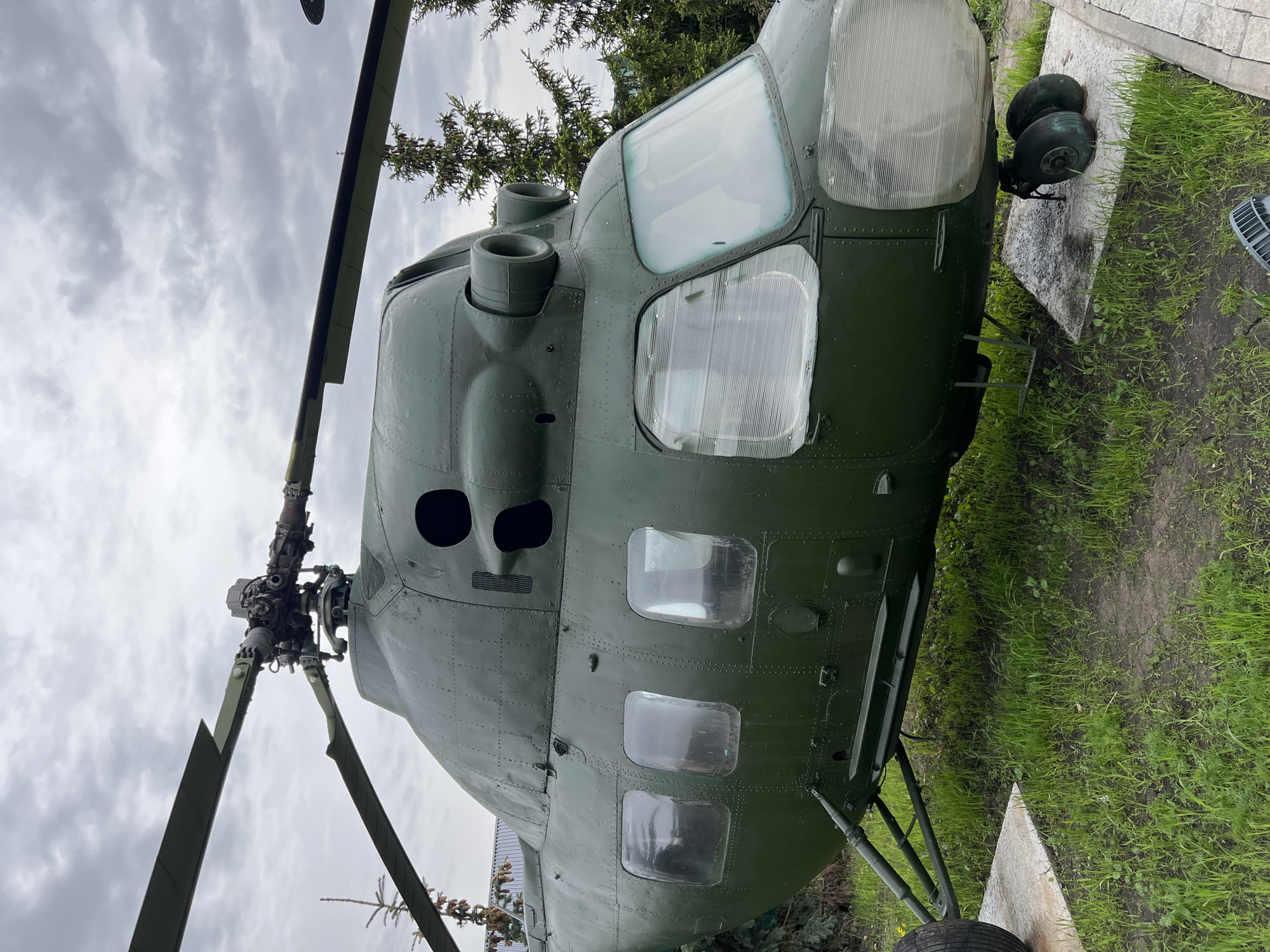 The image features a static display of a green Mil Mi-8 helicopter, likely in a park or outdoor museum setting. The helicopter's cockpit windows are prominent, and the main rotor blades are visible against a cloudy sky. The aircraft is situated on a concrete platform, surrounded by green grass and foliage. Given the mention of Введенская Слобода, Russia, it can be assumed that this display is located within or near that city.