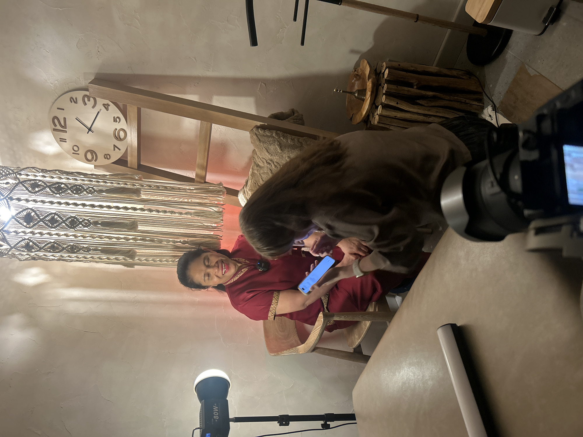 A woman wearing a red traditional garment is seated in a wooden chair and smiling while looking at a smartphone held by another person. The setting appears to be indoors, possibly a studio or a decorated room. A large macrame tapestry hangs on the wall behind the seated woman, and a wooden ladder with a clock is positioned to her right. A professional light is set up to the left of the scene, and a camera is visible in the foreground, suggesting a recording or filming session. The floor has some scattered items, including a piece of cardboard and a cable. The overall ambiance is warm and inviting, with soft lighting. There is no indication of the location being in Postol, Russia.