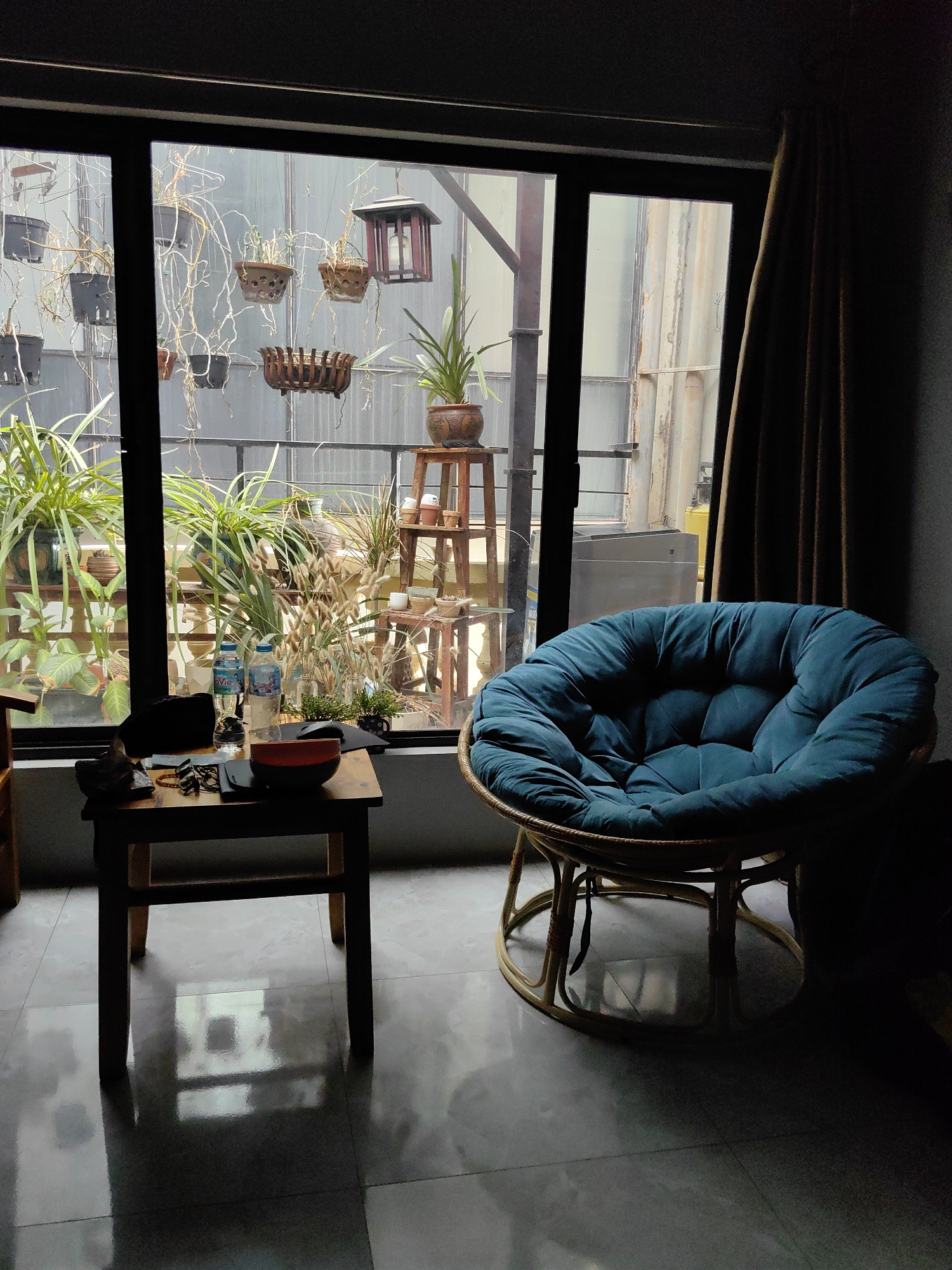 The image shows an interior scene in Hà Nội, Vietnam, evidenced by the water bottles on the small table bearing the “Lavie” brand, a bottled water brand commonly available in Vietnam. The scene is set near a window that offers a view of an exterior space, possibly a courtyard or balcony, populated with various plants, both potted and hanging. The window frame is dark, contrasting with the daylight visible outside. A small wooden table stands to the left, bearing several items, including a bowl, a wallet, a pair of glasses, and the aforementioned water bottles. To the right of the window is a large, round, cushioned chair in a blue-green color, partially obscured by the room's shadow. A curtain is draped to the right of the window, adding to the dimness of the interior. The floor appears to be tiled and reflects light from the window. Outside, there is a mix of vegetation and what appears to be a metal structure or wall, suggesting an urban residential setting.