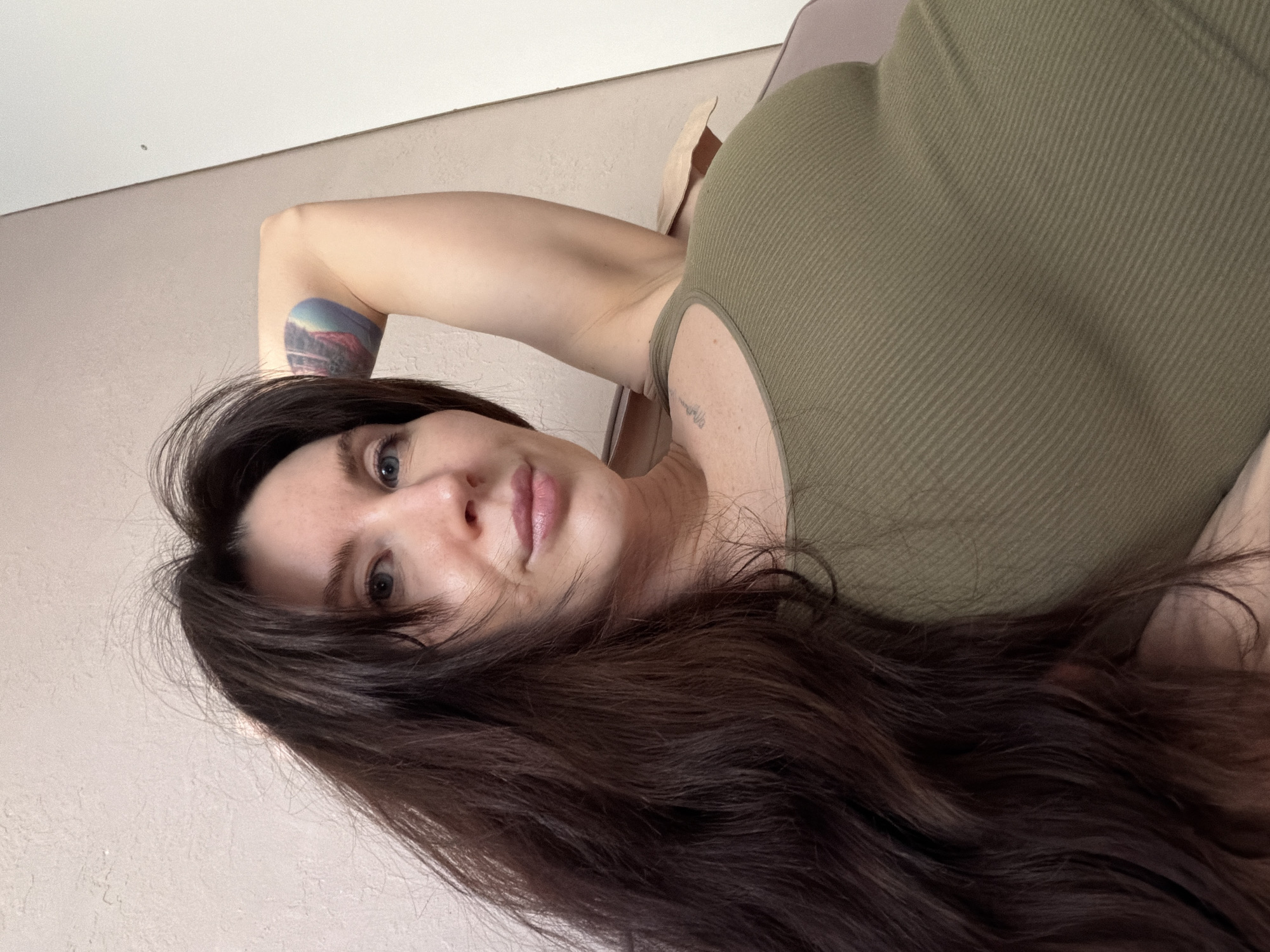 A woman with long, dark brown hair is pictured in a close-up shot. She has fair skin, blue eyes, and is wearing a ribbed, olive-green tank top. A tattoo is visible on her left arm, and another smaller tattoo is near her collarbone. The background is a light, neutral-colored wall. The photograph does not depict a scene in Perm, Russia and contains no details related to this city.