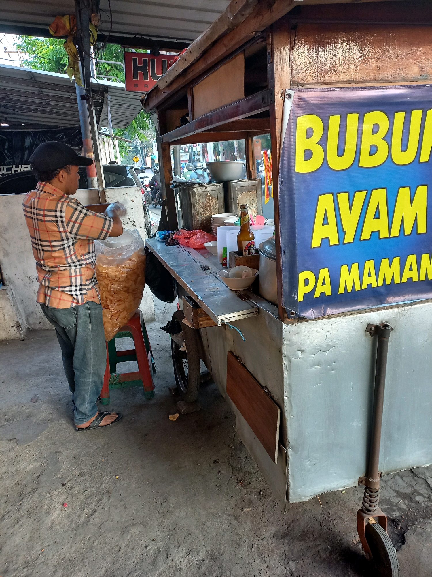 A man wearing a plaid shirt and jeans stands behind a food cart, holding a large plastic bag filled with chips. The food cart has a banner with Indonesian text that appears to say "BUBUR AYAM PA MAMAM" which translates to "Chicken Porridge Eat". Several items are on the cart's counter, including bowls, bottles, and a red plastic stool. The scene appears to be outdoors, with a street and trees visible in the background. The ground is a dirt and concrete mix. This setting is typical of street food vendors often found in cities like Cimahi, Indonesia.