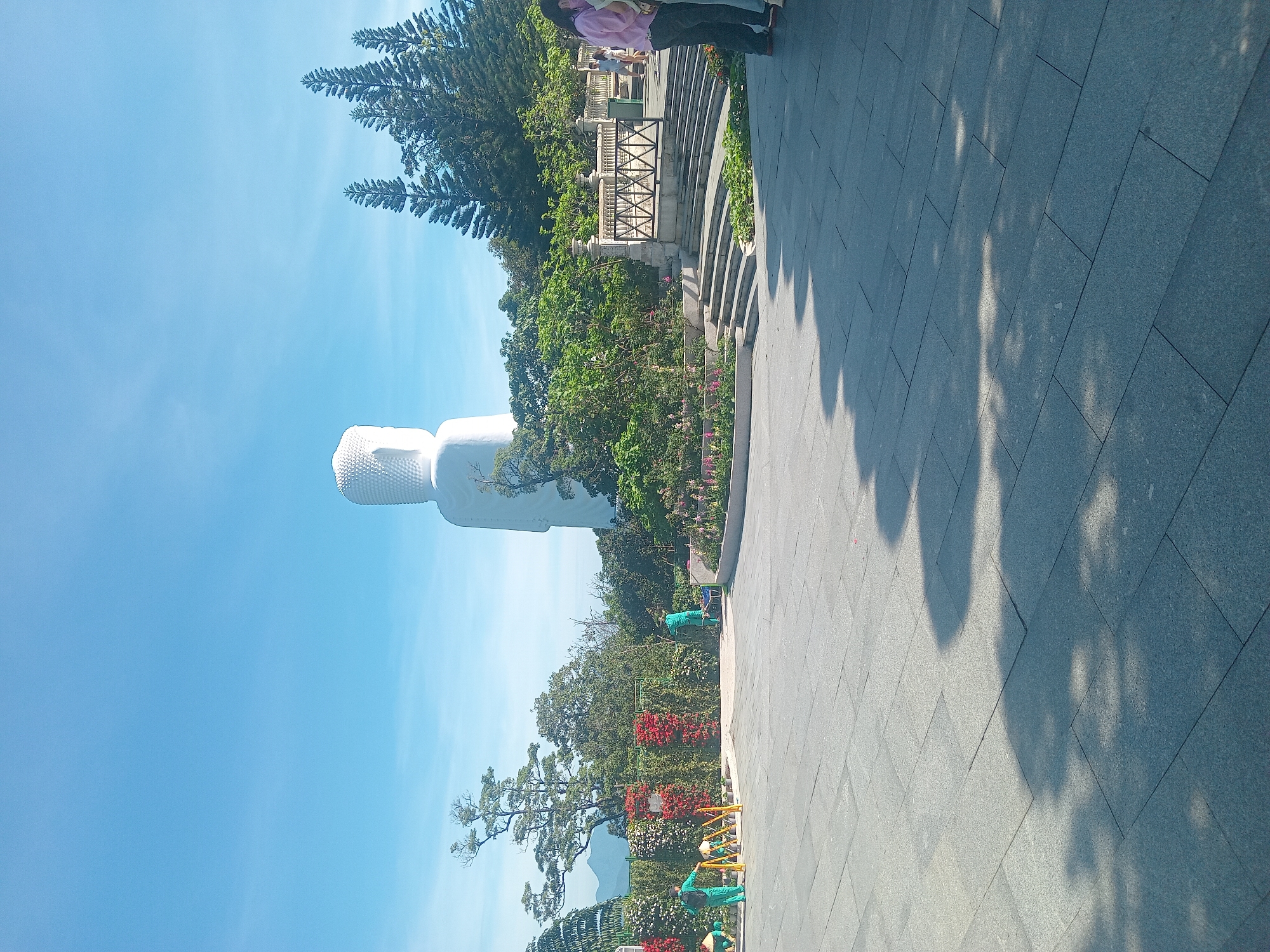 This image depicts the Linh Ung Pagoda on Son Tra Peninsula in Da Nang, Vietnam. A colossal white Buddha statue stands prominently against a clear blue sky, its head and upper body visible above lush greenery. The statue is draped in white fabric, hinting at its construction or repair. In the foreground, a paved plaza is laid with large grey stone tiles, casting dappled shadows from unseen trees. To the left, gardeners in green uniforms are tending to colorful flower beds and bushes. Further back, a series of tiered stone steps lead up to a balustrade. Several visitors are visible near the steps, including a woman in a pink shirt and dark pants and another in a light blue shirt. The overall scene is serene and bathed in bright sunlight, suggesting a pleasant day for tourism and spiritual reflection.