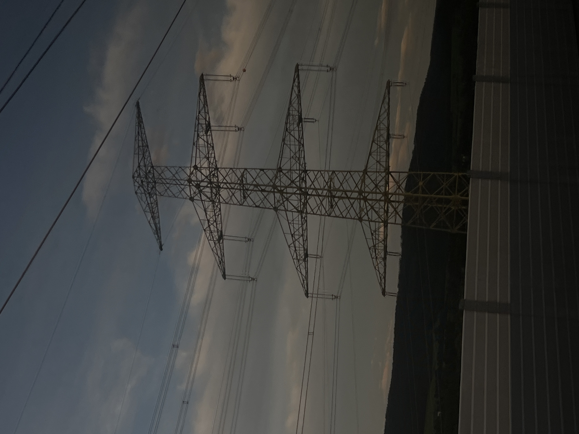 A towering electrical pylon, constructed of metal lattice, stands prominently against a twilight sky. Multiple power lines crisscross the frame, extending from the pylon and disappearing into the distance. In the mid-ground, a dark, undulating hillside forms the backdrop, with hints of greenery visible. In the foreground, a horizontally oriented, light-colored barrier, possibly a sound barrier or guardrail, runs across the bottom of the image, its textured surface suggesting a series of parallel ridges. The overall scene suggests a rural or semi-rural environment, possibly on the outskirts of Ilmenau, Germany, given the context. There are no people or animals visible in the image, and no discernable action or interaction is taking place. The atmosphere is one of quiet and solitude, with the fading light casting long shadows and imbuing the scene with a somber mood.