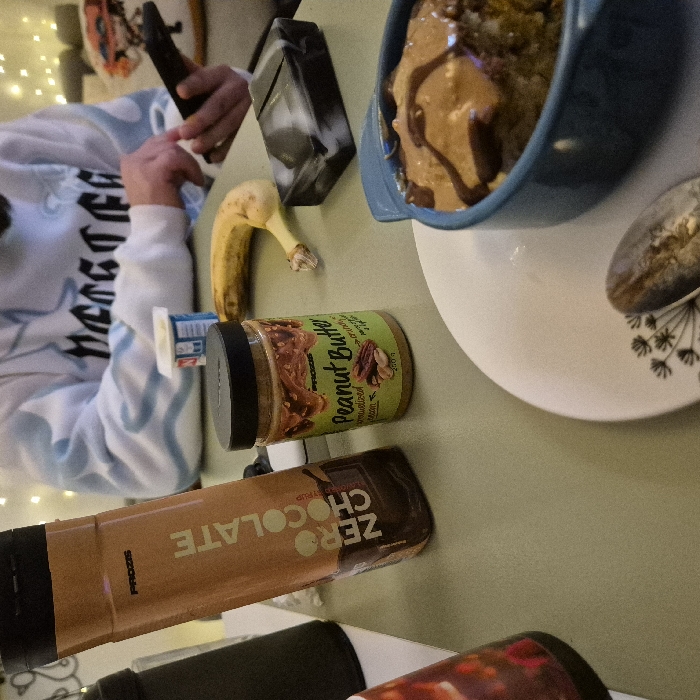 The image displays an indoor setting, likely a casual dining area, with a light green table surface prominently featured. On the table, a tall brown bottle labeled "PROZIS ZERO CHOCOLATE SYRUP" stands upright to the left. Adjacent to it on the right is a smaller jar of "PROZIS Peanut Butter crunchy caramelized pecan" with a black lid. Further right, a single ripe banana lies horizontally. A black and white marbled rectangular object is situated to the right of the banana. In the bottom right foreground, a blue ceramic ramekin contains a dessert, possibly a baked good or oatmeal, topped with a lighter brown substance and a dark brown drizzle. This ramekin rests on a white plate adorned with a black floral pattern, alongside a silver spoon. In the background, a person wearing a light-colored sweatshirt with dark lettering and light blue wave patterns is visible, holding a black smartphone in their right hand. A small white yogurt container with a blue and red label is visible near the person's left hand. Out-of-focus warm white string lights are visible in the upper background. No discernible visual cues within the image specify a particular location in the city of Strasbourg, France.