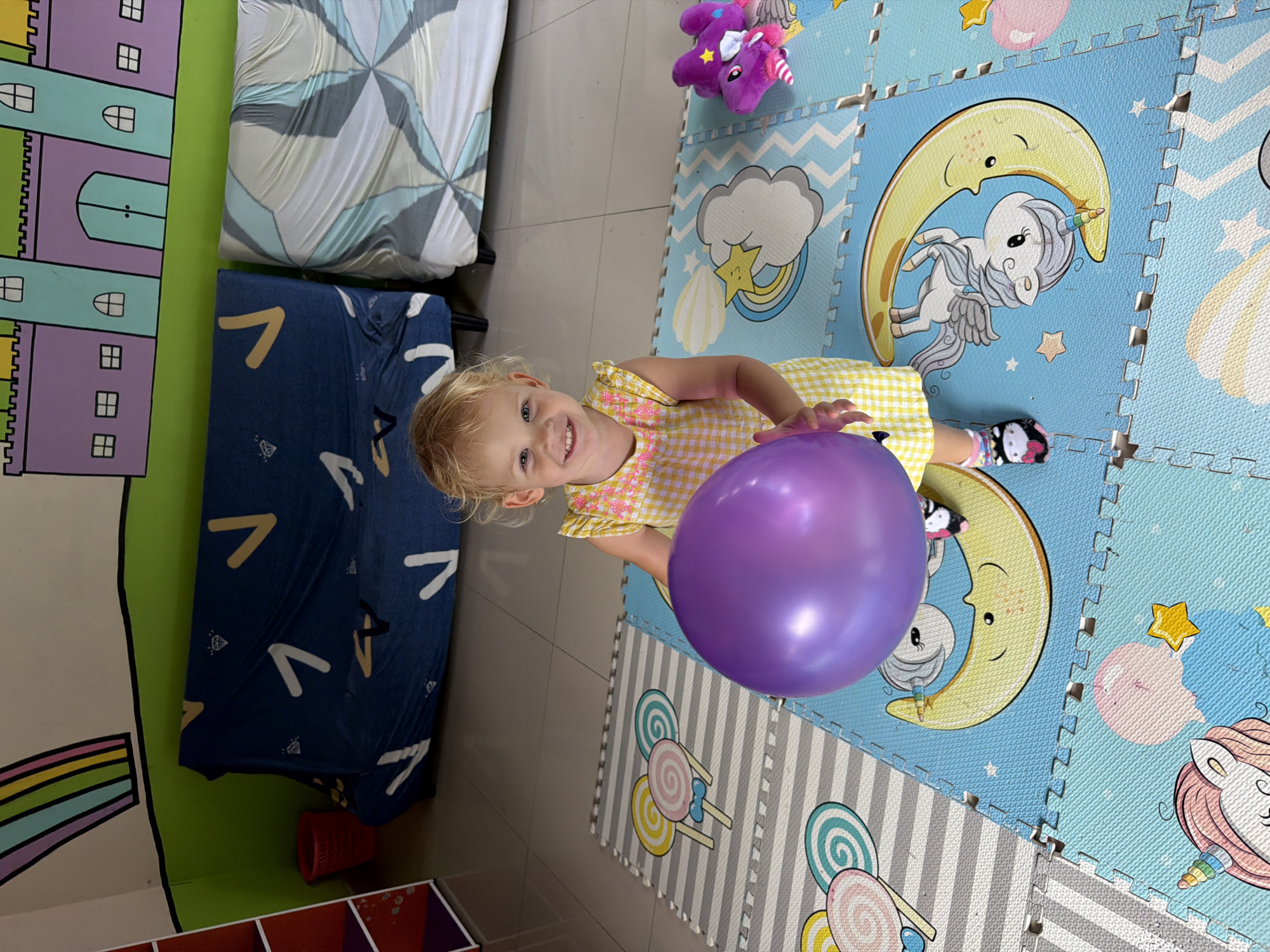 A young Caucasian girl with blonde hair and blue eyes is holding a large purple balloon. She is wearing a yellow and white checkered dress. The girl is lying down on a soft mat that has a playful design of moons, clouds, and unicorns. In the background, there is a blue blanket with yellow and white patterns, a green wall with colorful castle drawings, and a larger white and grey mat. A small purple stuffed unicorn toy is visible near the top of the frame. The scene appears to be indoors, possibly in a child's play area. The lighting suggests it is daytime. The girl has a happy and engaged expression, looking directly at the camera. The location is Kuta, Indonesia. There is no visible text in the image.
