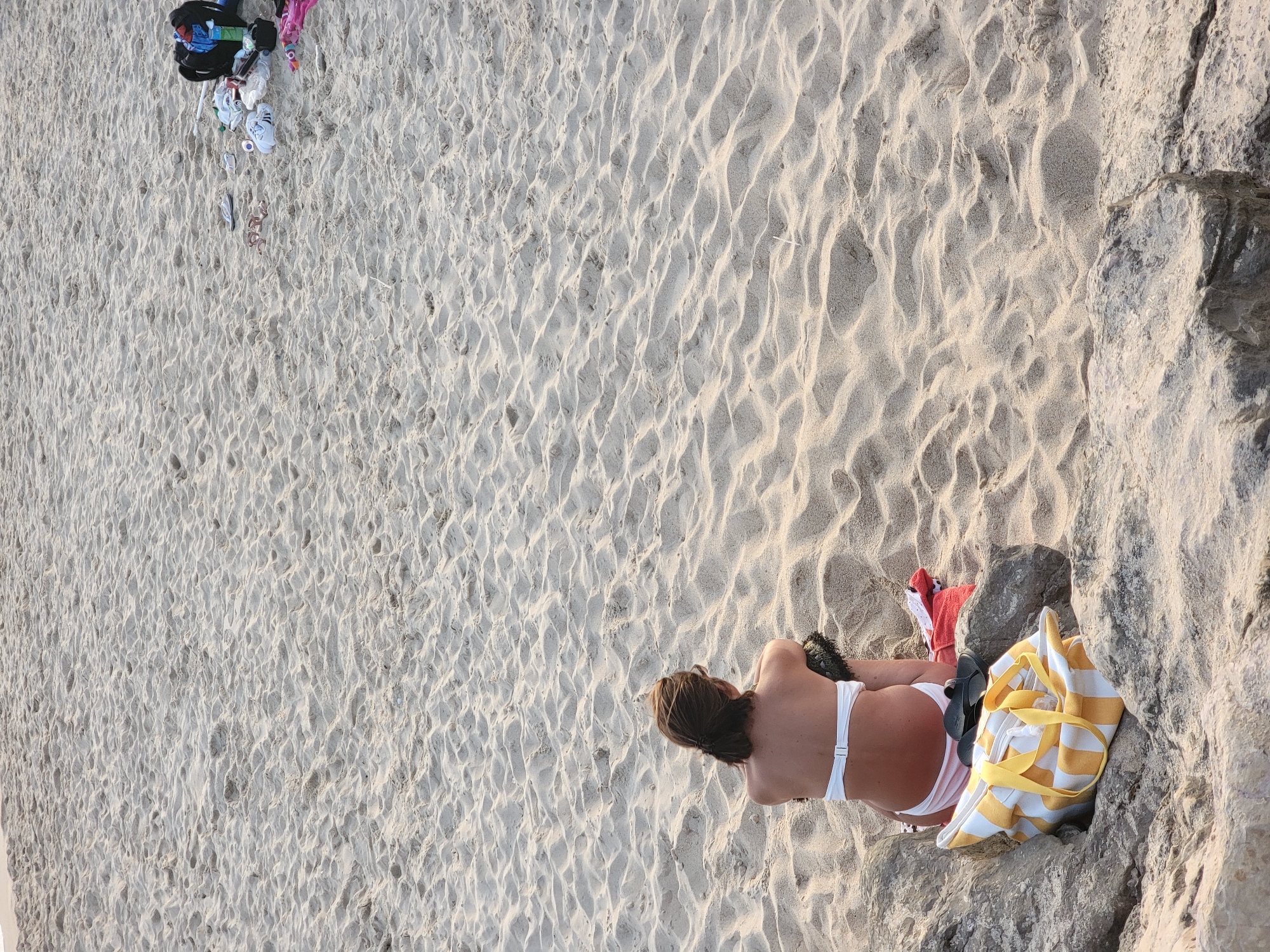 A woman in a white bikini sits on a sandy beach in Quarteira, Portugal, facing away from the camera. She is positioned on a rocky outcrop, with a yellow and white striped beach bag at her feet. Further up the beach, a collection of items including a black backpack, sandals, and other beach accessories are scattered on the sand. The sand is rippled, indicating the passage of wind or water, and the overall scene depicts a typical day at the beach.