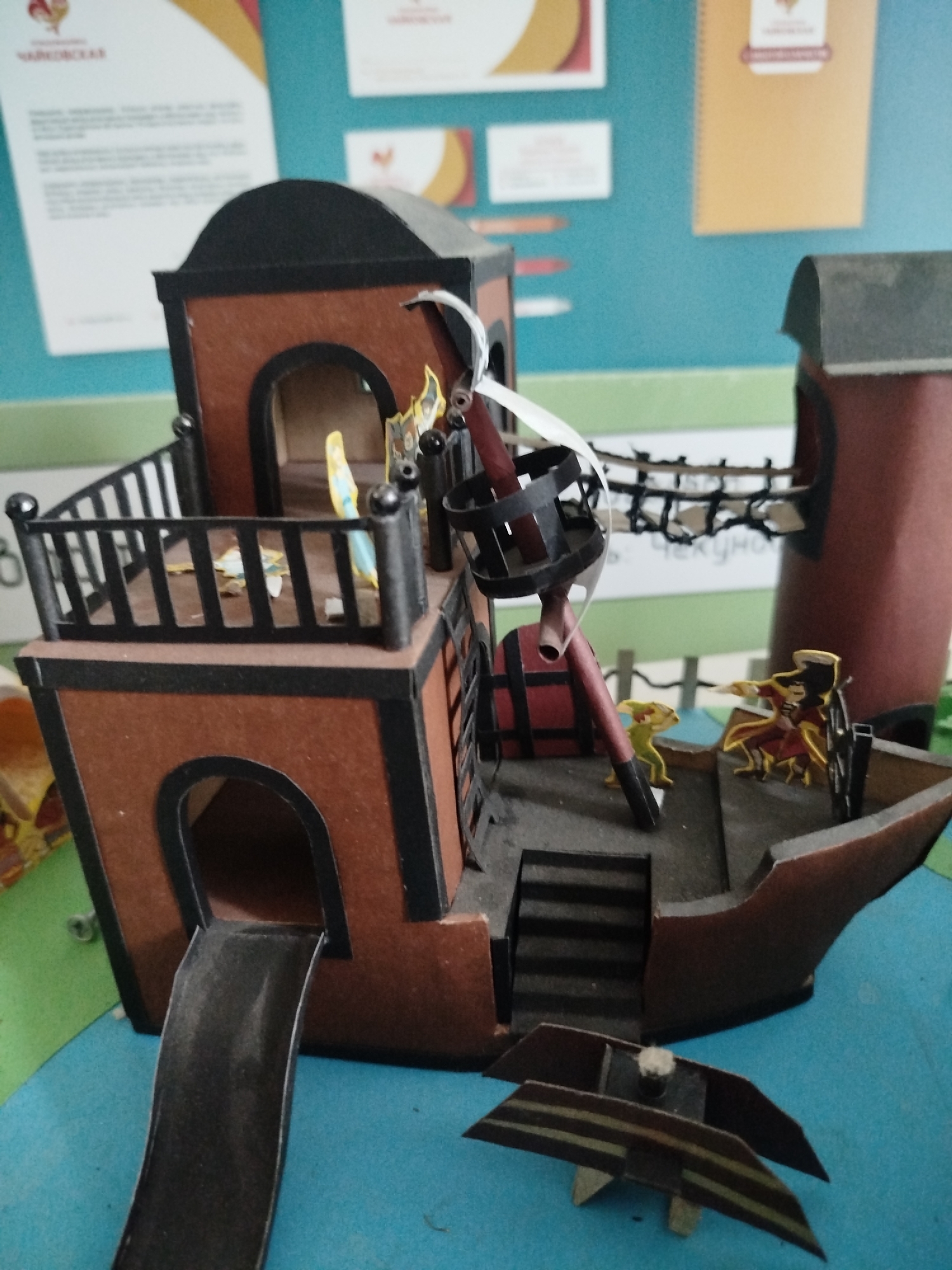 A cardboard model of a pirate ship sits on a blue surface, possibly a table or floor. The ship is brown and black, with a deck, railings, a mast with a basket, and a slide. Two small figures, one yellow and one red and black, are on the deck, appearing to be in conversation or conflict. The background shows a teal wall with posters and other papers attached. There are no visible signs of the city or any specific location.