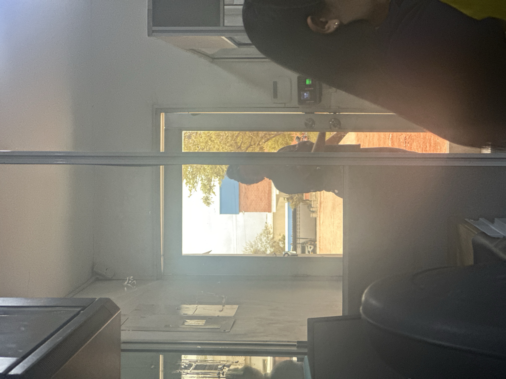 The image depicts an indoor scene, likely an office or administrative building, with a person silhouetted in the doorway leading outside. The doorway reveals an outdoor environment bathed in bright daylight, with trees and buildings visible. The interior is dimly lit and cluttered with office furniture, including chairs and what appears to be a computer monitor in the foreground. On the wall near the doorway, a security camera is visible. There is also a digital lock system mounted on the wall next to the door. A portion of a woman's face and hair is visible on the right side of the frame, suggesting she is seated inside. The overall impression is one of an active but somewhat confined workspace. The location is identified as Juan Mina, Colombia.