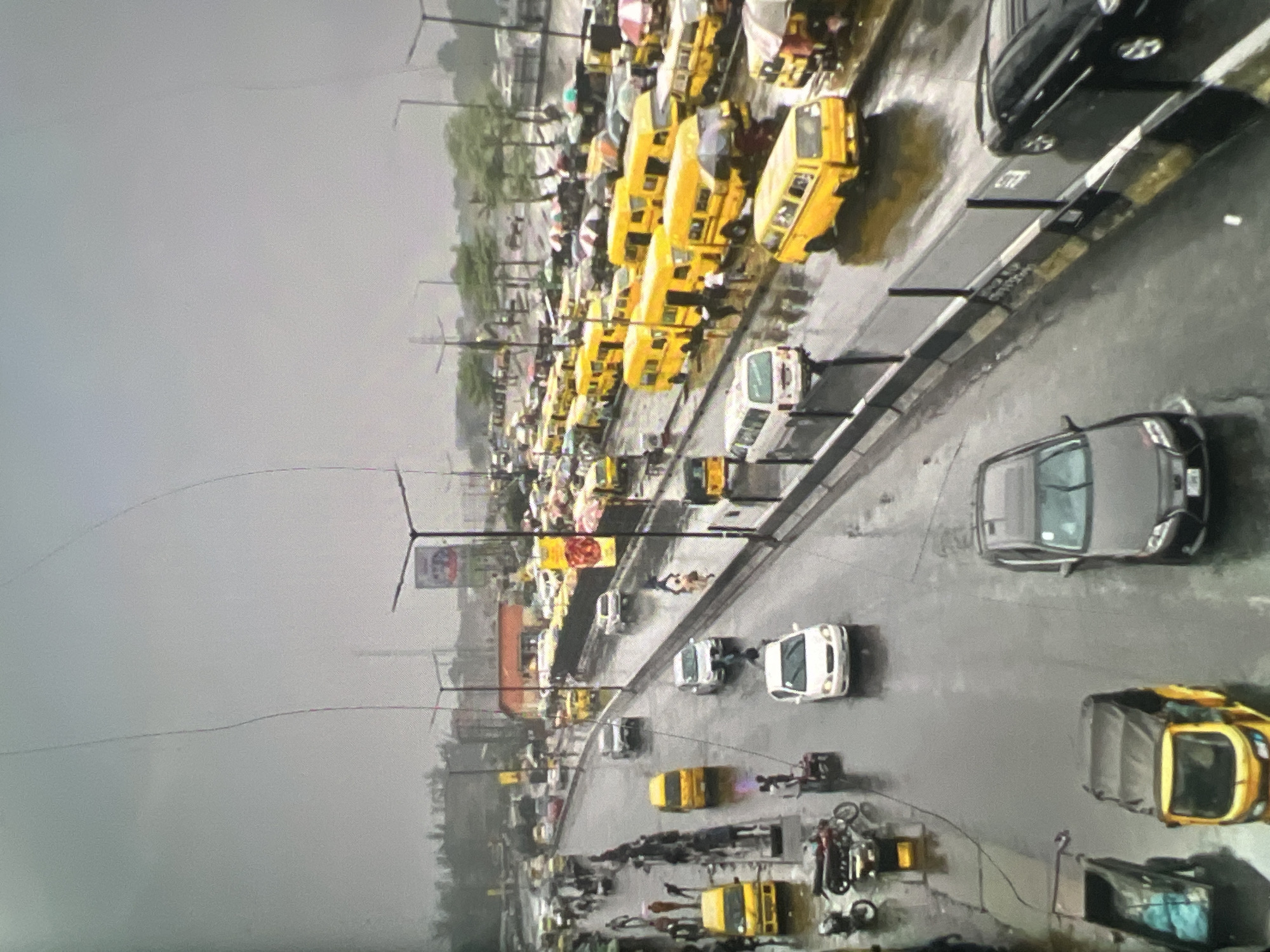 The image depicts a rainy day in a busy urban area, likely Lagos, Nigeria, characterized by numerous yellow buses, cars, motorcycles, and people on a wet road. The scene is an overhead perspective of a street with multiple lanes of traffic. The weather is overcast, with visible streaks across the sky that may be rain or camera artifacts.
Key elements in the scene include:
* **Vehicles:** A multitude of yellow buses, many of which appear to be minibuses or "danfos" typical of Lagos public transport, are parked and moving. Several cars of various colors (white, silver, black) are also present, as are motorcycles and a three-wheeled auto-rickshaw. Some vehicles have their headlights on.
* **People:** Numerous individuals are visible walking on the sidewalks and crossing the road. Some are holding umbrellas, indicating the rain. Pedestrians are dressed in everyday clothing.
* **Environment:** The road surface is wet and reflective, indicating rainfall. There are sidewalks lined with trees. Buildings and billboards are visible in the background, suggesting a commercial or densely populated urban setting. Streetlights are also present.
* **Activity:** The scene conveys a sense of bustling activity despite the rain. Vehicles are navigating the traffic, and pedestrians are moving about their daily routines. The presence of many buses suggests a major transportation hub or route.
The overall impression is one of a vibrant city life continuing under challenging weather conditions. The yellow buses are a distinctive visual element, strongly suggesting the location as Lagos. The presence of market stalls with umbrellas in the background further points to a busy commercial area.