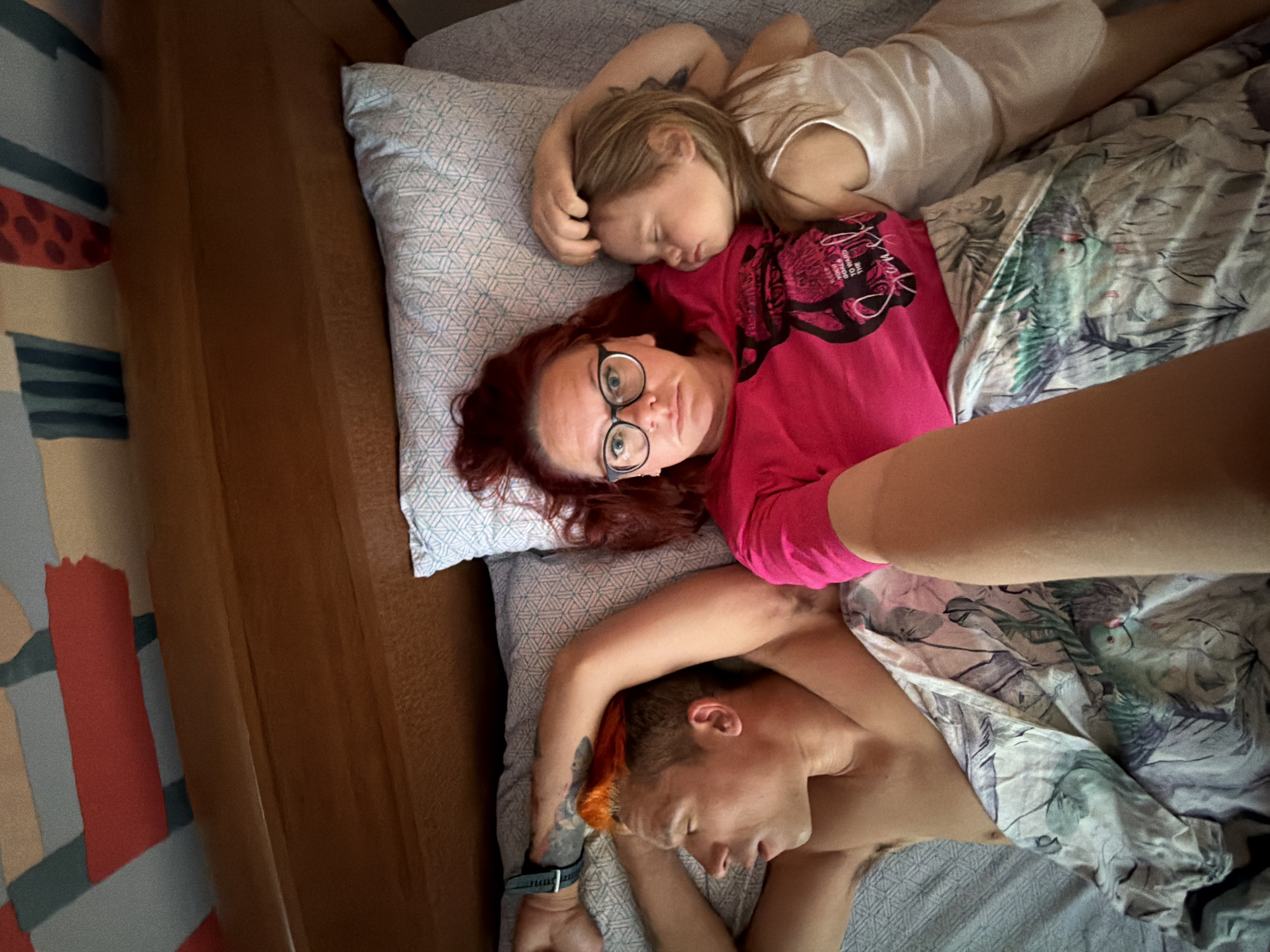 The image shows three individuals resting together in a bed, viewed from an overhead perspective. An adult woman with red hair and black-rimmed glasses lies awake on a pillow, looking directly at the camera with a neutral expression. She is wearing a bright pink t-shirt that displays partially visible black text, including "THE PINK PANTHERS WILL" and other smaller words.
Resting on her upper body is a young child with blonde hair, fast asleep in a white top. Below the red-haired woman, another adult with short, orange-dyed hair is visible, lying on their back, also appearing to be asleep or resting. This individual is bare-chested and has a dark watch on their left wrist, along with visible tattoos on their arm. One of their arms is gently draped over the red-haired woman's shoulder.
The bed features light-colored pillows with a subtle geometric pattern. A duvet covers the lower half of the people, displaying a distinctive light green and blue tropical leaf and bird pattern. A wooden headboard or bed frame is visible along the left side of the image, and further left, a patterned curtain or wall hanging with blocks of red, teal, and beige colors can be seen. The scene conveys a moment of familial intimacy and rest, likely indoors during the daytime in a bedroom in Chaikovsky, Russia.