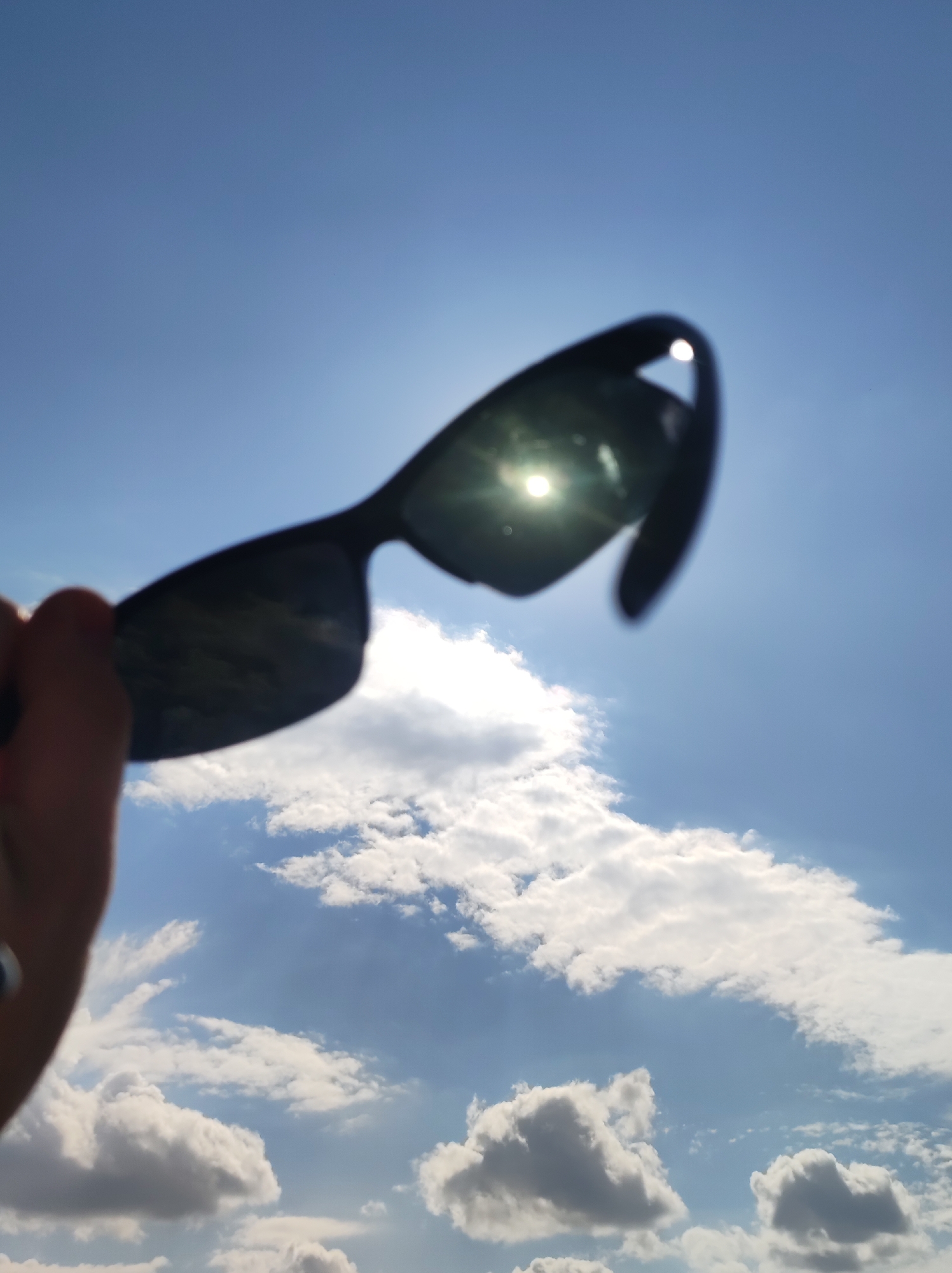 The image displays a bright, daytime sky over what is contextually identified as Фоки, Russia. In the foreground, a person's hand is visible on the left, holding a pair of dark-framed sunglasses upwards. The sunglasses are slightly out of focus, with the sun's intense glare reflecting prominently off their dark lenses, creating a bright starburst effect. The sky is a clear, vibrant blue, punctuated by numerous white and light grey cumulus clouds of varying sizes and shapes. Some clouds are fluffy and isolated, while others form larger, more elongated masses. The overall scene suggests a sunny day with scattered clouds. The perspective is from a low angle, looking up towards the sky and the hand holding the sunglasses. No other specific objects, people, or text are visible within the frame.