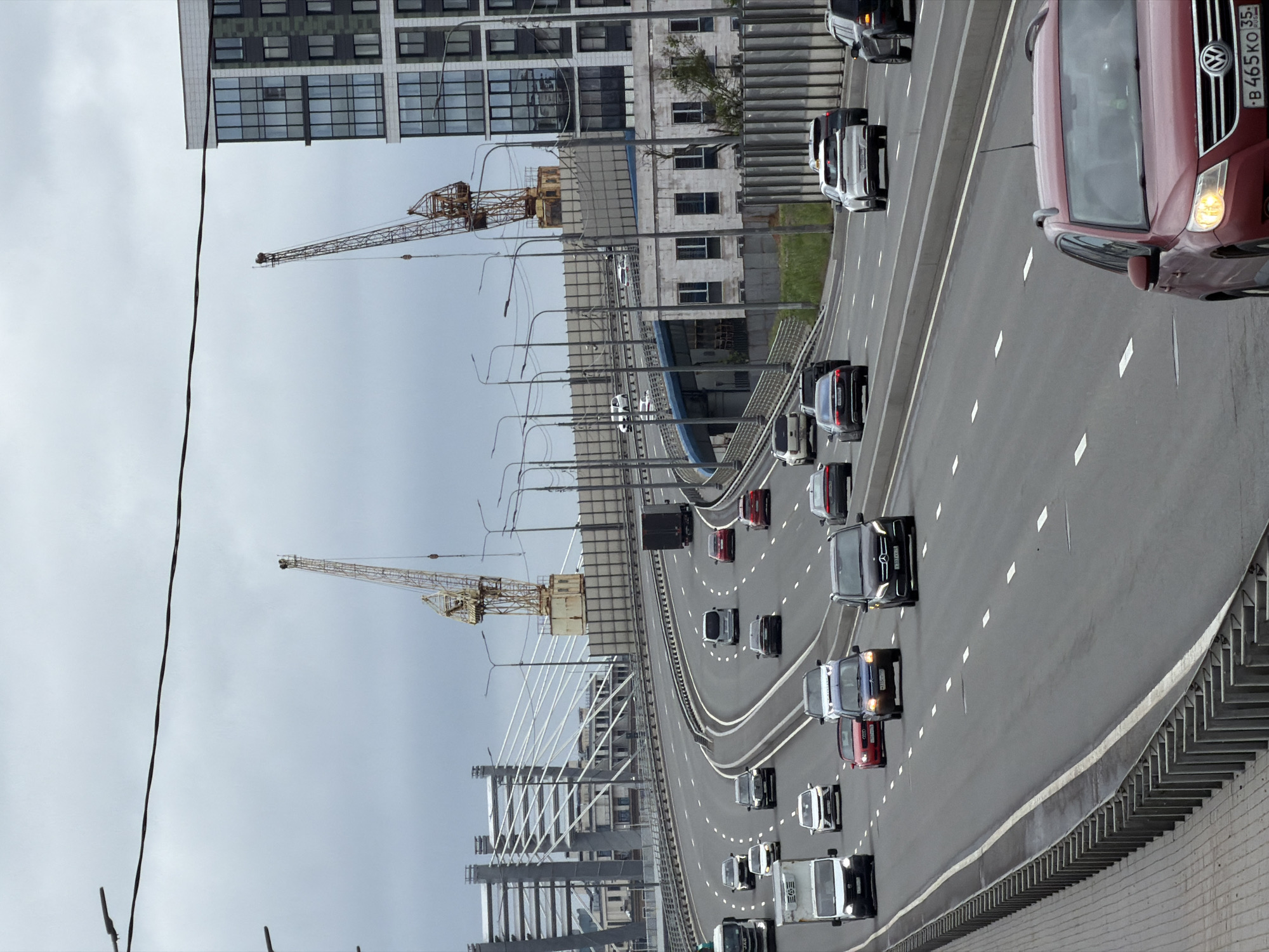 The image shows a view of the Western High-Speed Diameter (WHSD) highway in Saint Petersburg, Russia. The highway is elevated and features multiple lanes of traffic in both directions. Cars, trucks, and vans are visible on the road, with various makes and models. In the background, there are large modern buildings, including a tall, multi-story structure with a glass facade. Two large construction cranes are also prominent in the background, suggesting ongoing development or construction in the area. The sky is overcast, indicating a cloudy day. The WHSD is a major toll road that bypasses the city center and connects different parts of Saint Petersburg. The scene captures a typical urban transportation environment with vehicular traffic and architectural elements characteristic of a developed city.