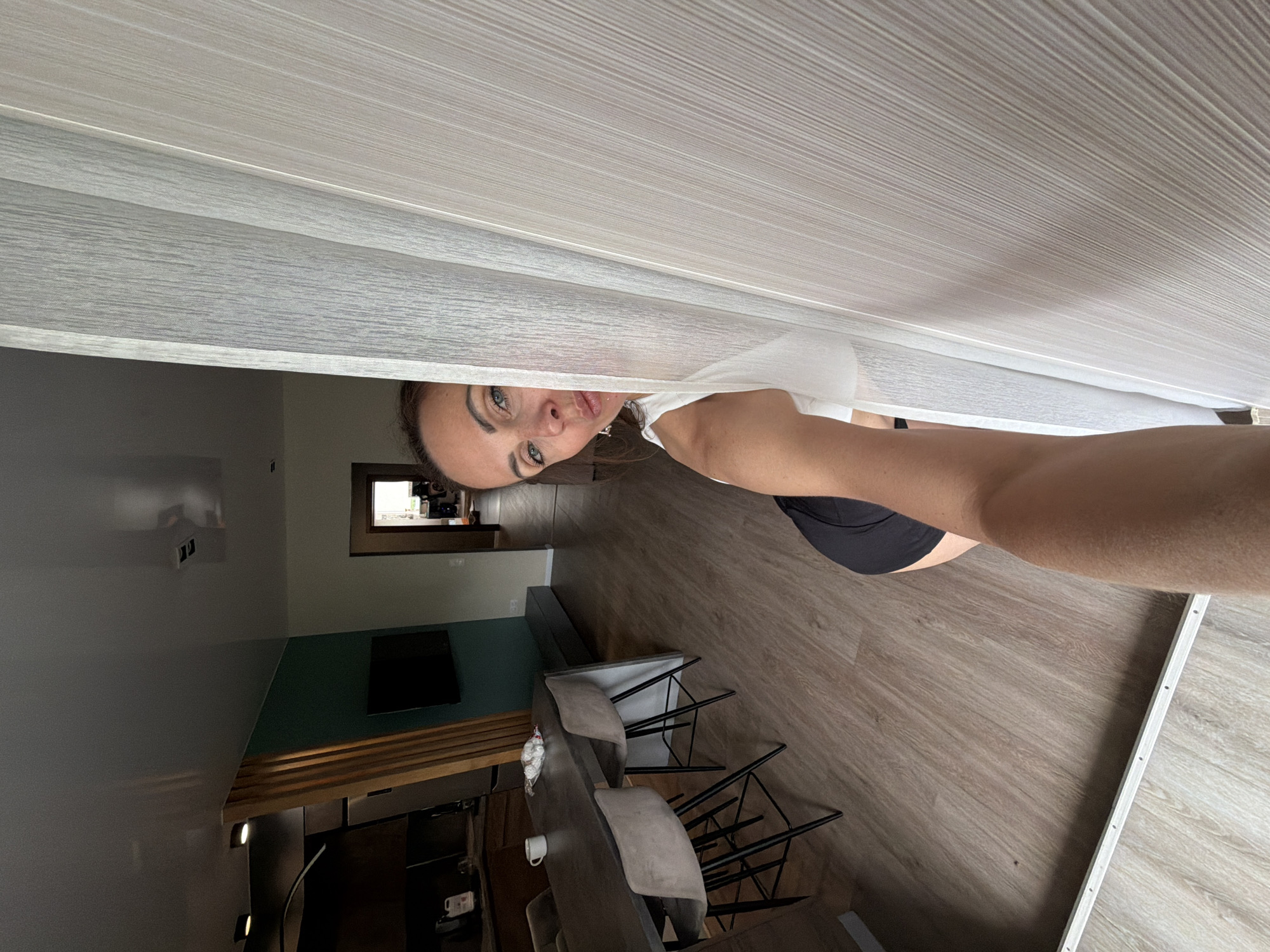 The image shows a woman peeking from behind a vertical white blind in a modern apartment. She is wearing a white top and black shorts, with her right arm extended towards the camera. Her face is partially visible, with her eyes looking directly at the viewer and her lips slightly parted. The background reveals a dining area with a dark table and chairs, a kitchen counter with a mug and a bag of marshmallows, and a television mounted on a teal wall. The flooring is light-colored wood laminate. A doorway in the background leads to another room, where some gym equipment is visible. The scene is indoors, suggesting a residential setting.