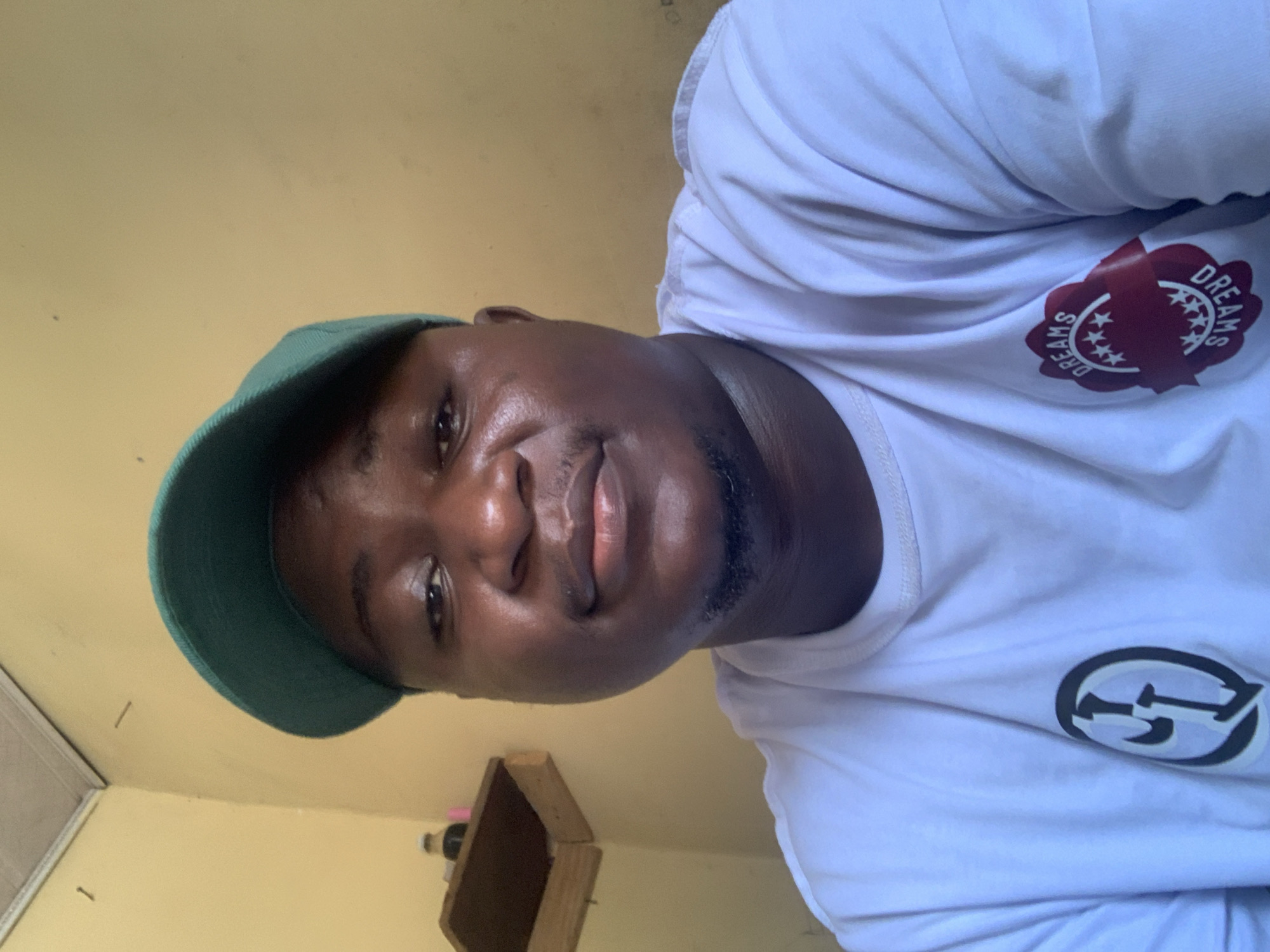 The image is a close-up selfie of a young man, likely taken indoors in Akwanga, Nigeria. The man has dark skin, short dark hair, and a neatly trimmed beard. He is wearing a dark green baseball cap and a white short-sleeved t-shirt. His expression is neutral to slightly serious as he looks directly into the camera.
His white t-shirt features two distinct logos. On the upper right side (viewer's left), there is a red circular emblem with the word "DREAMS" written in white, encircling three white stars in the center. On the lower left side (viewer's right), a black and white circular logo with a stylized letter or symbol, possibly a 'G', is visible.
The background consists of plain, light yellow-colored walls. To the bottom right of the frame, a simple wooden box or shelf is present, holding a small bottle with a red cap and a white cylindrical object. A few small dark specks and a nail are also visible on the wall. The lighting suggests the image was taken during the daytime.
