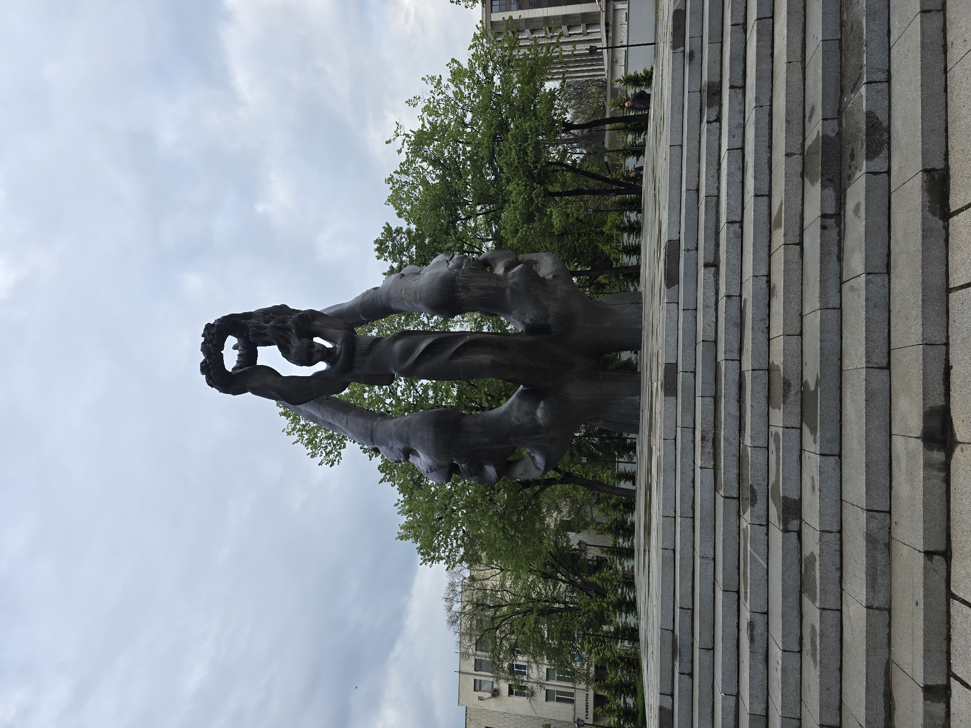 Here is a forensic description of the image:
The image shows a bronze monument in a city park. The monument depicts several figures intertwined, with one figure holding up a wreath. The monument is set against a backdrop of trees and buildings. The monument is surrounded by stone steps. The sky is overcast. The monument is located in an unknown city.