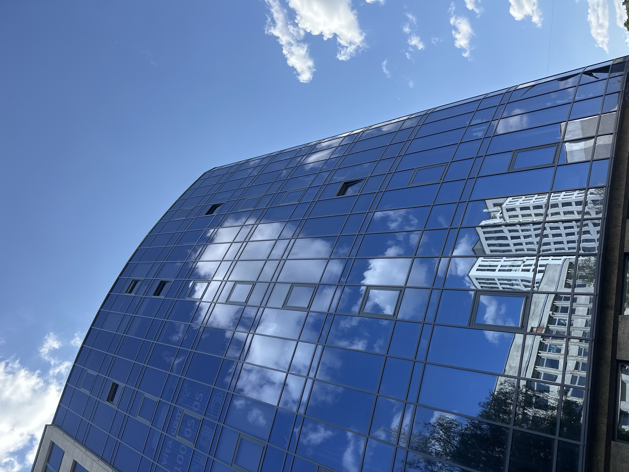 The image shows a modern glass-facade office building in Ternopil, Ukraine, with a clear blue sky and scattered white clouds above. The building's reflective glass mirrors the sky, clouds, and an adjacent older building. The older building appears to be a light-colored structure with multiple windows. The glass facade of the modern building is segmented by dark frames, and some windows are open. Text is visible on some of the glass panels, though it is partially obscured and difficult to read clearly. The perspective is from a low angle, looking up at the imposing structure. The overall impression is one of urban architecture under a bright, sunny sky.