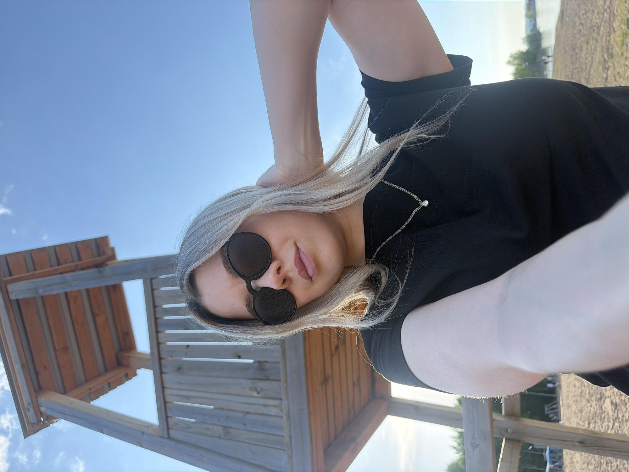 A selfie shows a woman with blonde hair wearing round, black sunglasses, a black shirt, and a silver necklace with a clear gemstone pendant. She is standing next to a wooden lifeguard tower. The sky is blue, and in the background, there is a body of water. The location is Chaikovsky, Russia.