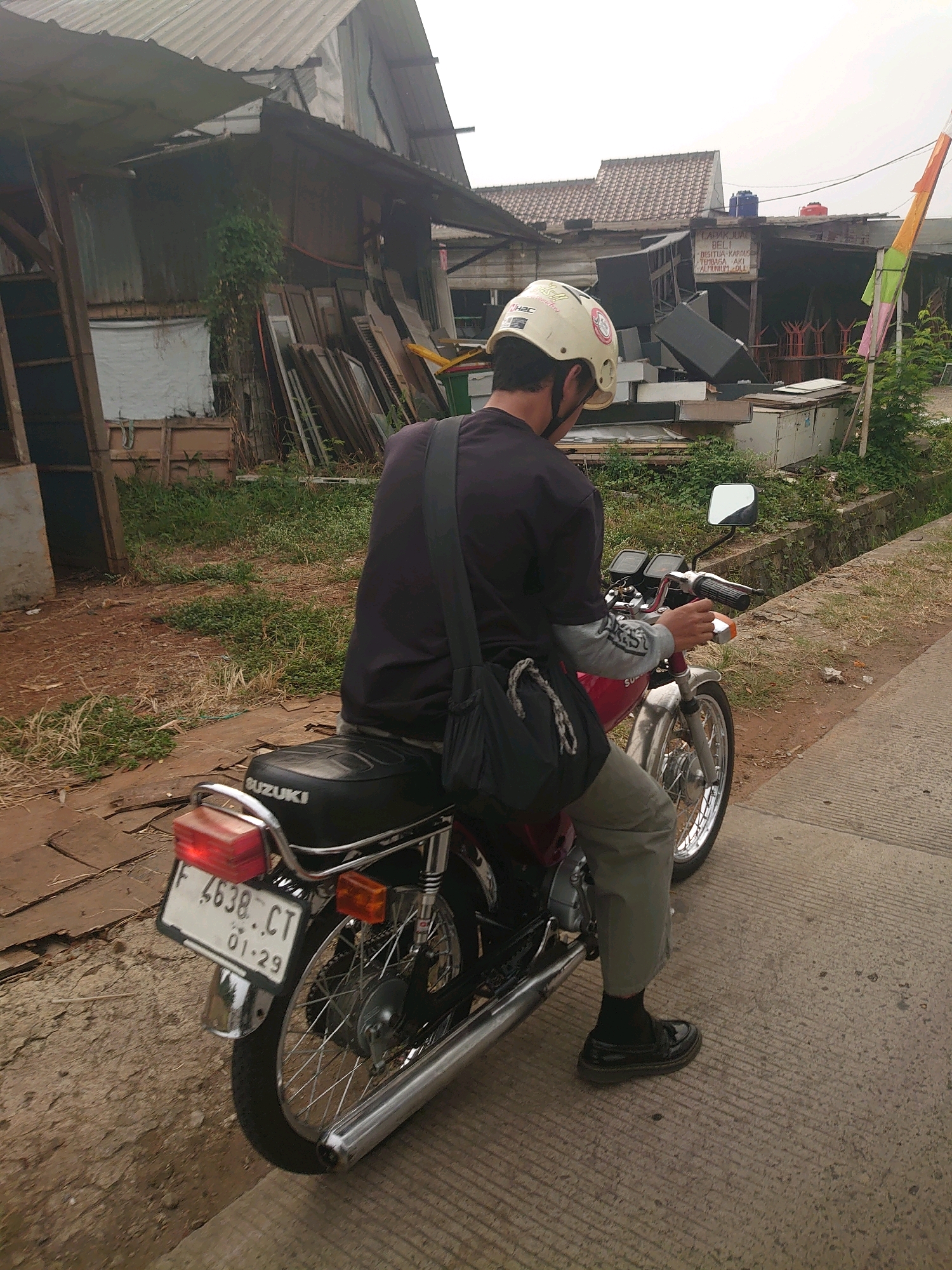 A person wearing a white helmet, a dark purple shirt, and light gray pants sits on a red and black Suzuki motorcycle. The motorcycle has a license plate that reads "F 4638 CT 01-29". The person is looking away from the camera. The background shows a dilapidated building with a corrugated metal roof and wooden walls, stacks of wood, and overgrown vegetation. There are also some plastic chairs and what appear to be speakers stacked up. A colorful flag is visible on the right side of the image. The scene appears to be in a somewhat neglected urban or semi-urban area. Based on the license plate format, this is likely Depok, Indonesia.