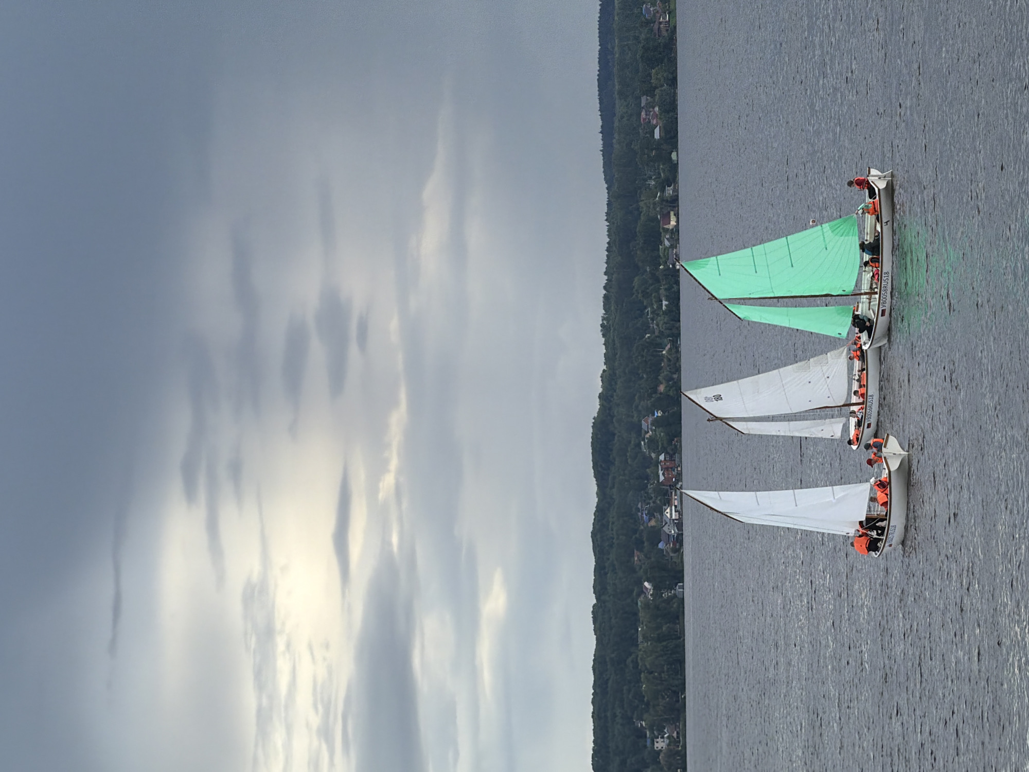 The image captures a scene of three small sailboats, likely rowing dinghies equipped with sails, moving across a body of water near Votkinsk, Russia, under an overcast sky. The boats are positioned diagonally from the bottom-left to the upper-right if the image were properly oriented.
Each boat carries multiple individuals, all visibly wearing bright orange life jackets for safety. The vessel closest to the viewer (bottom-left if upright) has a white, triangular sail. Its hull bears the alphanumeric text "VB0057RU618". Behind it, another boat features a striking bright green sail and the hull inscription "VB0058RU618". Further back, partially obscured, is a third boat with a white sail, on which the number "808" is visible.
The water surface is calm with subtle ripples, suggesting gentle conditions for sailing. In the background, a tree-covered shoreline extends along the horizon, dotted with numerous houses and structures, indicating a settled area. The sky is predominantly grey with soft, diffused light filtering through dense clouds, indicating an overcast day. The activity appears to be a organized sailing event or training session, given the uniformity of the boats, the presence of multiple participants in each, and the safety equipment worn.