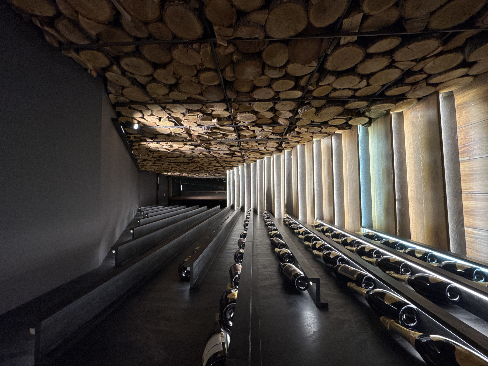 The image depicts an interior wine cellar in Blagoevgrad, Bulgaria. The ceiling is decorated with numerous circular wood slices. Wine bottles are displayed on tiered, black horizontal ledges along both walls of the narrow space. The right wall is accented with vertical, light-colored panels illuminated from behind, while the left wall is a flat, dark grey. Small, recessed lights are visible along the left wall. The floor is dark and reflective.