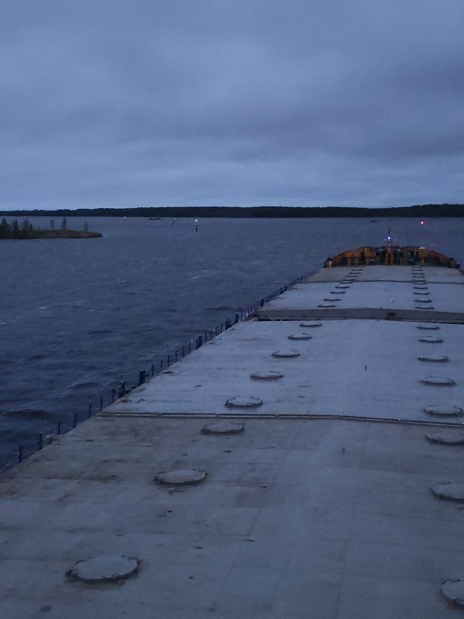 The image shows a section of a large, flat concrete structure, likely a barge or a dock, extending into a body of water. The water appears choppy and is bordered by land with trees in the distance. The sky is overcast and gray. The surface of the concrete structure has a grid-like pattern and several circular access points or manholes. A railing runs along the edge of the structure. In the distance on the water, there appear to be navigational markers or lights. The image was likely taken near Bereg, Russia, as per the request. No people are visible in the image.