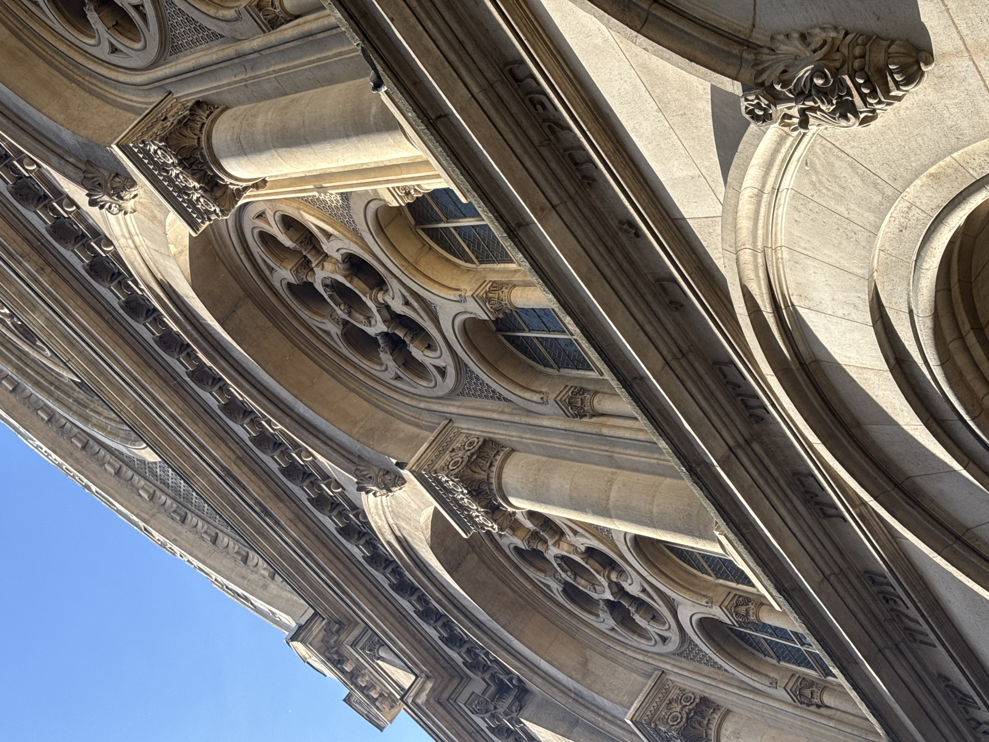This image presents a low-angle view of an intricately detailed stone building, likely located in Paris, France. The architectural style appears to be Gothic Revival or Neo-Gothic, characterized by ornate craftsmanship and decorative elements.
The primary subjects are the building's facade, composed of warm-toned, possibly limestone or granite, and a clear blue sky. No people or events are depicted.
Notable details include a series of deeply recessed arches, adorned with elaborate carvings and decorative friezes. Several circular windows, some featuring quatrefoil tracery, are visible, set within these arches. Columns topped with highly detailed capitals, sculpted with floral and foliate motifs, support various architectural layers. The intricate patterns extend to the spandrels between arches, showcasing fine masonry.
The bright sunlight casts strong, crisp shadows, highlighting the three-dimensional relief of the carvings and architectural projections. This suggests a clear, sunny day. The sky, visible in the lower left, is a vibrant, cloudless blue. The composition is dynamic, with the building's lines sweeping diagonally across the frame, emphasizing its grandeur and height. There is no visible text.