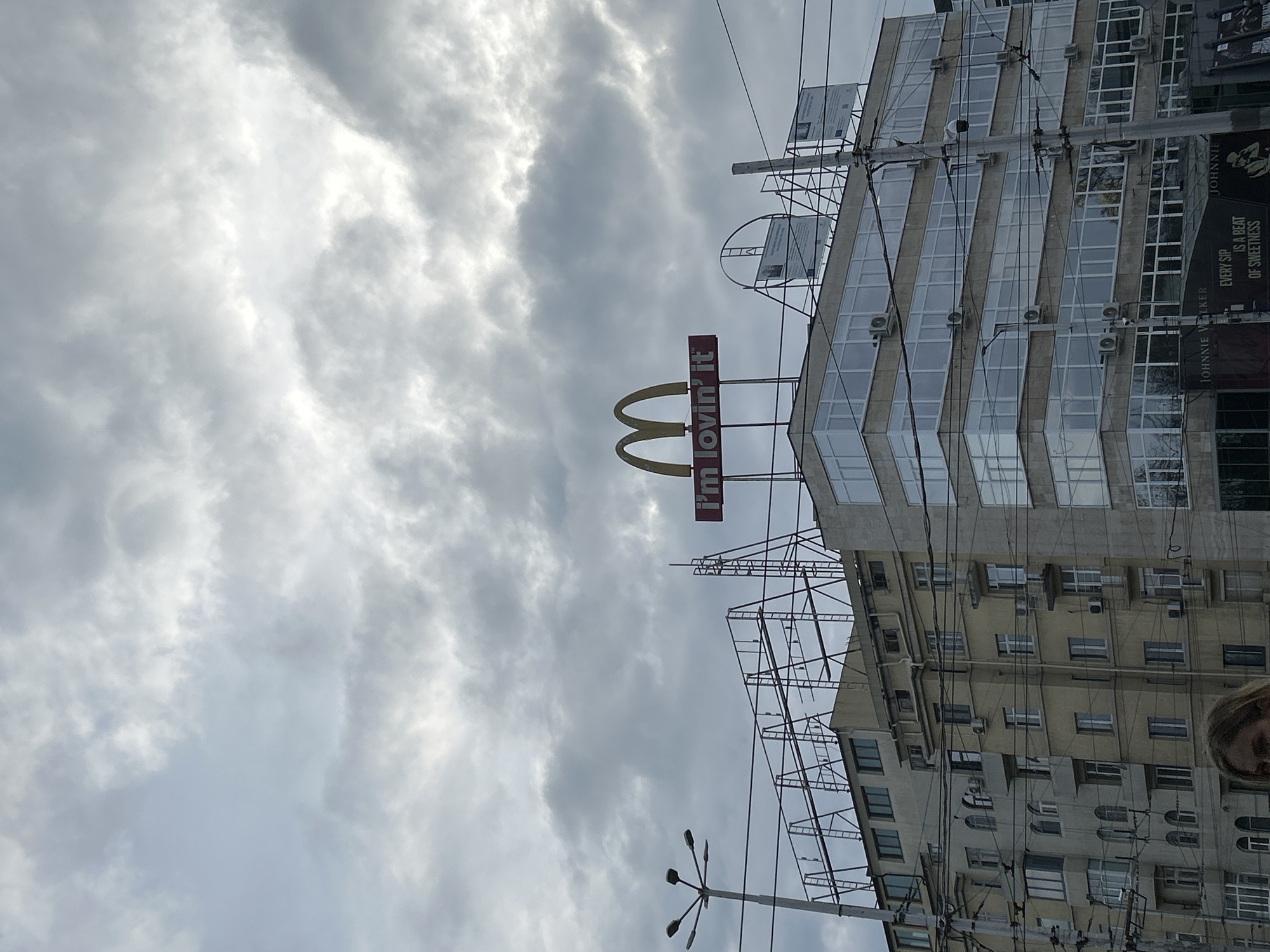 The image shows a McDonald's sign in Sofia, Bulgaria. The sign, featuring the golden arches and the slogan "i'm lovin' it," is mounted on top of a building. The building next to it appears to have a advertisement for Johnny Walker Whisky, specifically saying "EVERY SIP IS A BEAT OF SWEETNESS" The sky is overcast with heavy clouds. The building is a multi-story structure with numerous windows and air conditioning units mounted on the exterior. Power lines and poles are visible in the foreground. A person's face is slightly visible in the lower right corner of the image.