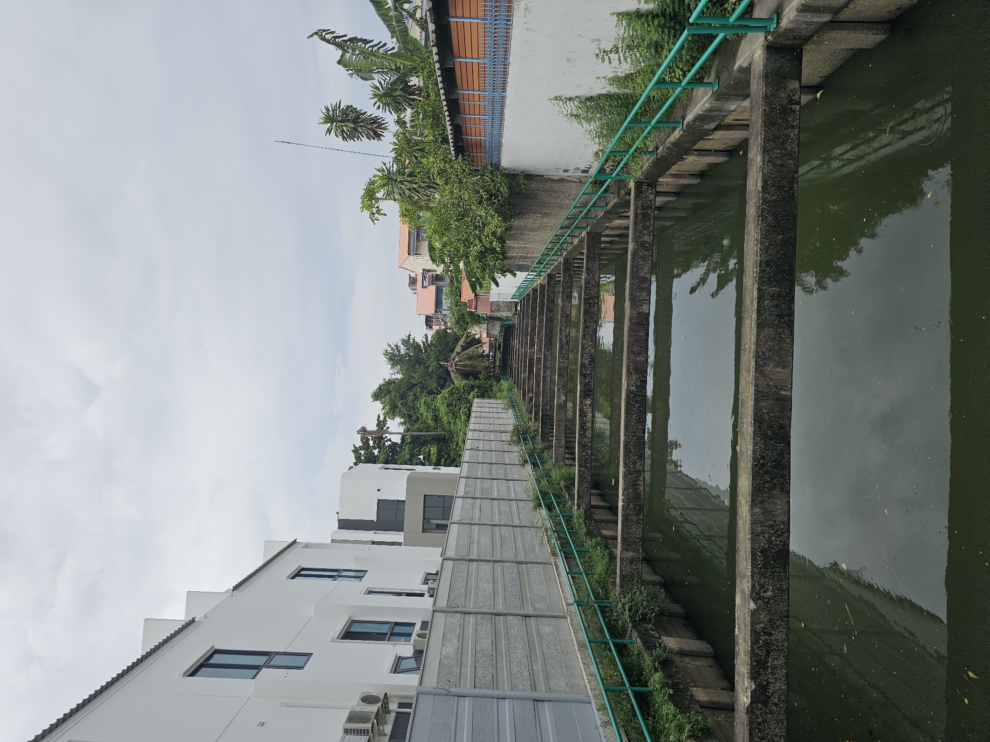 The image depicts a water channel with stepped concrete levels, characteristic of a water management system in Bangkok, Thailand. The channel is lined with green metal railings and surrounded by lush vegetation. On the left, a modern white building with large windows and air conditioning units is visible behind a concrete fence. On the right, a row of houses with terracotta roofs and wooden accents is seen beyond a white wall. The sky is overcast, suggesting a cloudy day. The water in the channel appears dark green and reflects the surrounding structures and sky. The scene captures a blend of urban infrastructure and natural elements typical of Bangkok's urban landscape.