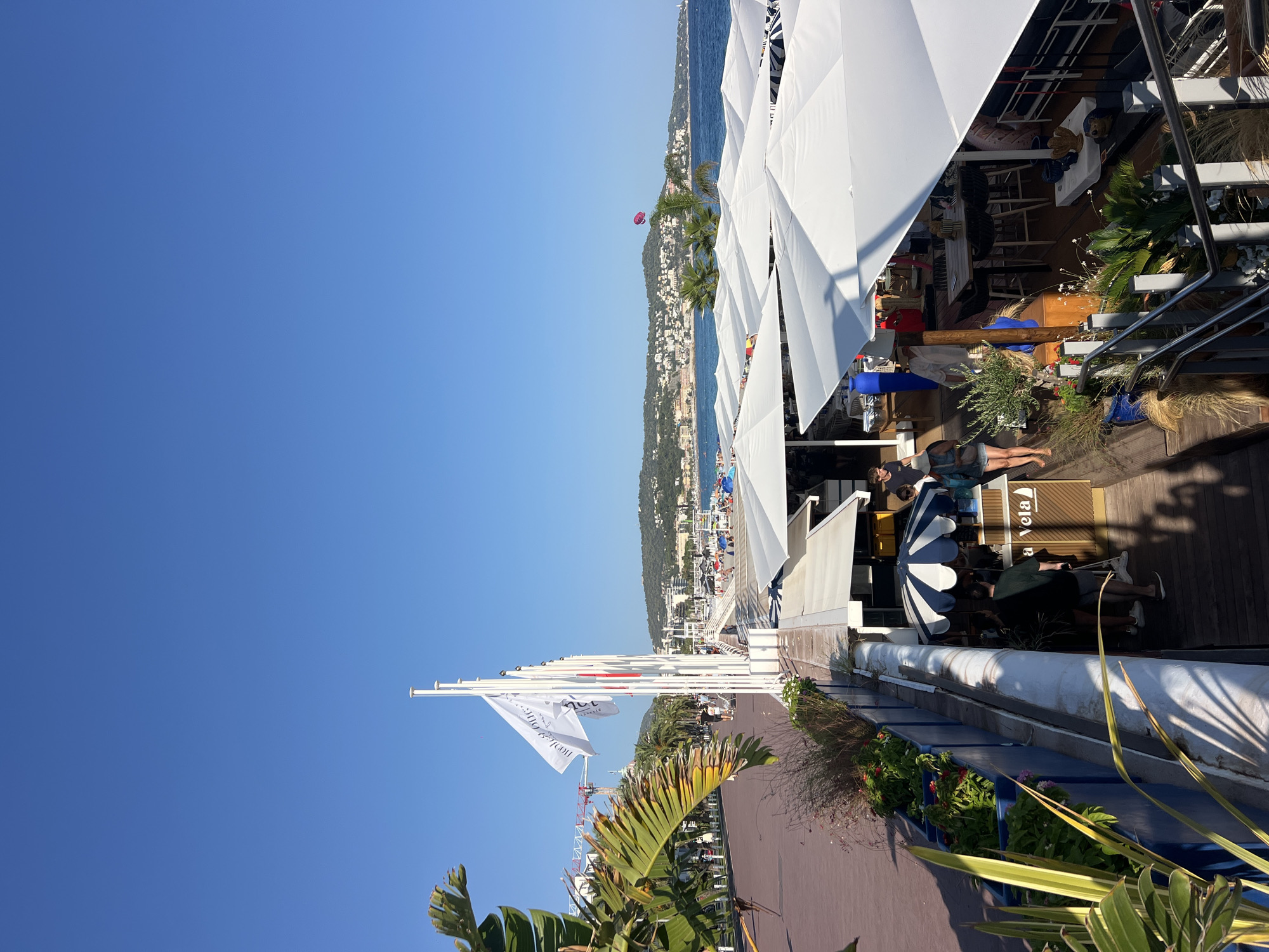The image captures a vibrant outdoor scene along the Promenade des Anglais in Nice, France, on a clear, sunny day. In the foreground, a walkway lined with palm trees and decorative plants leads towards a beachside establishment featuring numerous white umbrellas and outdoor seating areas. Several people are visible, some walking along the promenade, others seated at tables or browsing the establishment's offerings. The establishment appears to be a cafe or restaurant, with signage that includes the text "a Vela".
Further in the background, the iconic blue waters of the Mediterranean Sea stretch towards a picturesque coastline dotted with buildings and lush green hills. A parasailer is visible in the distance over the water. Flags fly on poles along the promenade, adding to the lively atmosphere. Construction equipment, specifically a crane, is visible in the far left background, suggesting ongoing development or maintenance in the city. The overall scene conveys a sense of leisure and enjoyment typical of a seaside resort.
