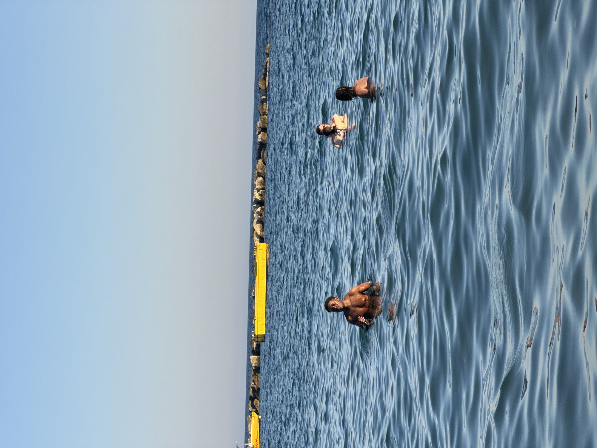 Four individuals are in the clear blue sea. In the foreground and to the left, a shirtless Black man stands in the water up to his chest, looking towards the left. Further back and to the right of him, two people are partially submerged in the water. One individual, a woman, is facing away from the camera, with her head and shoulders visible. The other, a person with dark hair, is facing towards the right, with their head and shoulders above water. In the background, a yellow float or buoy is visible, with rocks and a breakwater extending into the sea. Two yellow structures are visible in the far background. The sky above is clear and blue. The location is the city of Agios Tychona Community, Cyprus.