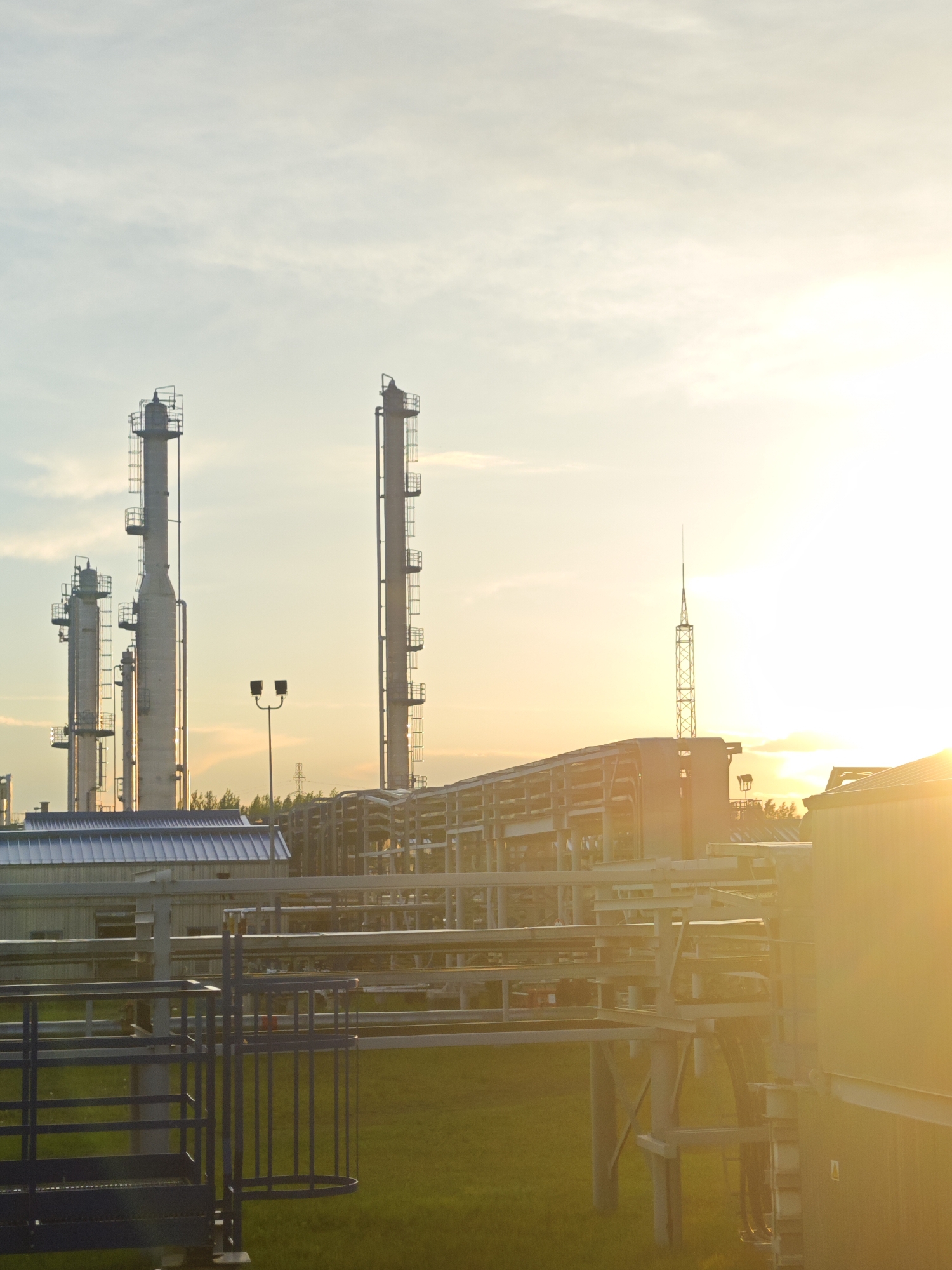This image depicts an industrial facility, likely an oil refinery or chemical plant, at either sunrise or sunset. The scene is dominated by several tall, cylindrical industrial columns, equipped with ladders and platforms, standing against a sky with scattered clouds. In the mid-ground, a streetlight with two lamps is visible, along with a building featuring a corrugated metal roof. To the right, the intensely bright, low sun creates significant lens flare and overexposure, silhouetting additional industrial structures, including multiple pipelines, support frameworks, and a tall, slender lattice tower with a pointed top. A large building or tank is partially visible on the far right, bathed in the bright sunlight. The foreground consists of a patch of green grass, partially illuminated by the sun, and a blue metal railing structure in the lower left. There are no people visible in the scene.