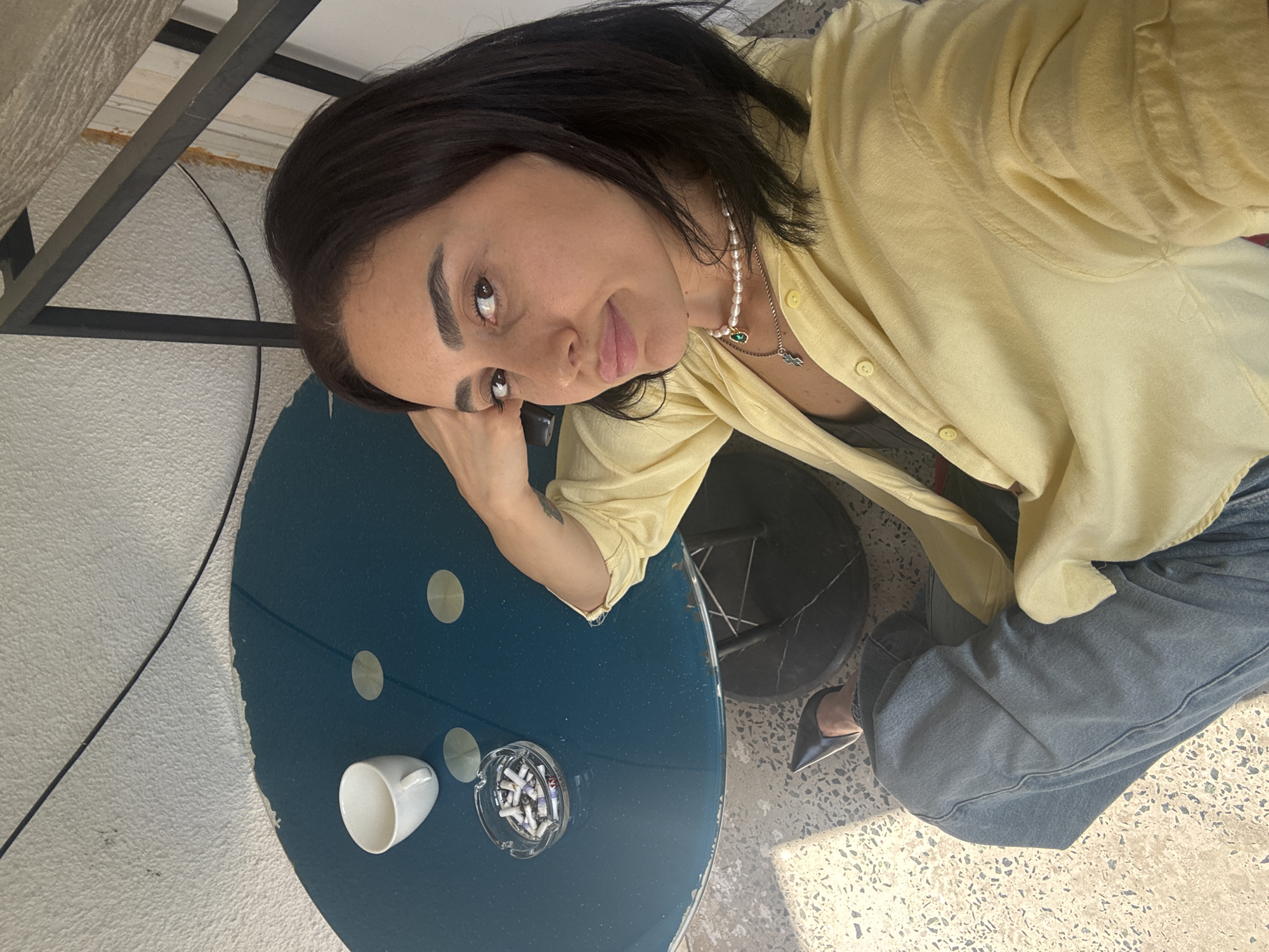 A woman is sitting at a round, teal-colored table, possibly outdoors on a patio or balcony. A white coffee cup sits on the table next to a glass ashtray filled with cigarette butts. The woman is wearing a light yellow button-up shirt, blue jeans, and black pointed-toe shoes. She has dark hair and is wearing a pearl necklace with a green pendant. Her right arm is resting on the back of a chair, and she is looking towards the camera. The background consists of a textured wall and a railing, suggesting an urban setting. The lighting indicates it is daytime. There is no specific indication of this being in Tbilisi, Georgia.