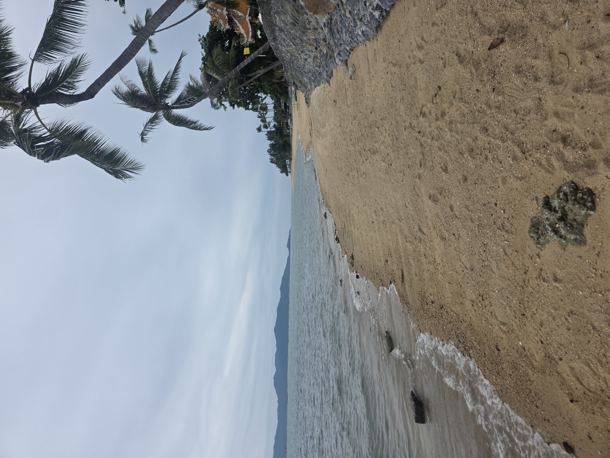 The image depicts a beach scene in Baan Bo Phut, Thailand, under a cloudy sky. The foreground is dominated by a wide expanse of light brown sand, with some small pebbles and a few scattered rocks. Gentle waves are lapping at the shore on the left side of the frame, with several larger rocks visible in the shallow water.
In the middle ground, the sandy beach curves to the left, lined with lush green palm trees and other vegetation. Further in the distance, a line of dark blue mountains stretches across the horizon. A partially visible structure with an orange and yellow tiled roof, possibly a temple or a resort building, is nestled among the trees on the right side of the beach. A yellow flag is also visible near this structure. The sea appears calm, with a muted blue-grey hue reflecting the overcast sky. The scene conveys a peaceful, natural ambiance of a tropical coastline. There are no people visible in the image.