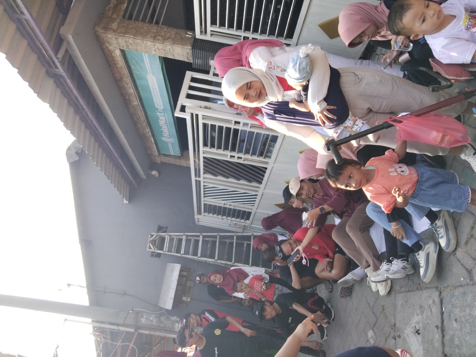 In the city of Kepuh Kiriman, Indonesia, a diverse group of people, including adults and children, are gathered outdoors on a paved area. The scene appears to be in front of a building with a gray facade and a corrugated metal roof, possibly a shop or commercial establishment given the "HalalMart" signage visible on a green awning above a white fence.
Several individuals are sitting on the ground, while others are standing. One woman in the foreground, wearing a white hijab and a light-colored outfit, is holding a baby. A stroller is visible near her. Among the seated individuals are men and women, some of whom are looking at their phones or interacting with each other. Children are also present, with one young boy in an orange shirt standing and looking towards the camera. A ladder is leaning against the building in the background. The lighting suggests it is daytime, possibly with bright sunlight casting shadows. The overall atmosphere appears casual and communal.