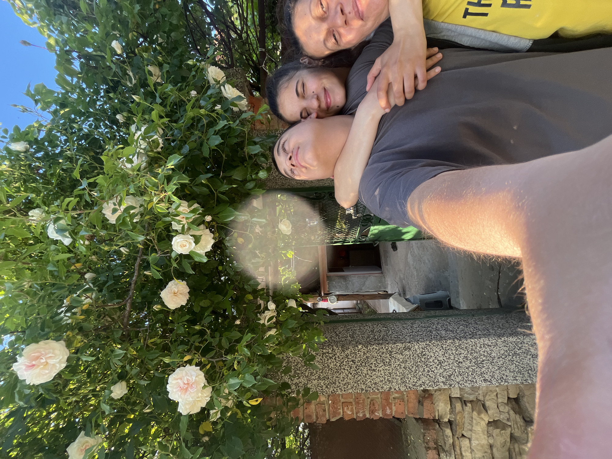 Three people are embraced closely, taking a selfie in an outdoor setting. A woman wearing a gray shirt hugs a younger person in a darker gray shirt. A third person, wearing a yellow t-shirt, is visible above the woman in the gray shirt. To the left, a flowering shrub with white blossoms dominates the frame. Behind the people, there are elements of a building, including a grey countertop and a brick structure. The location is Lozarevo, Bulgaria.