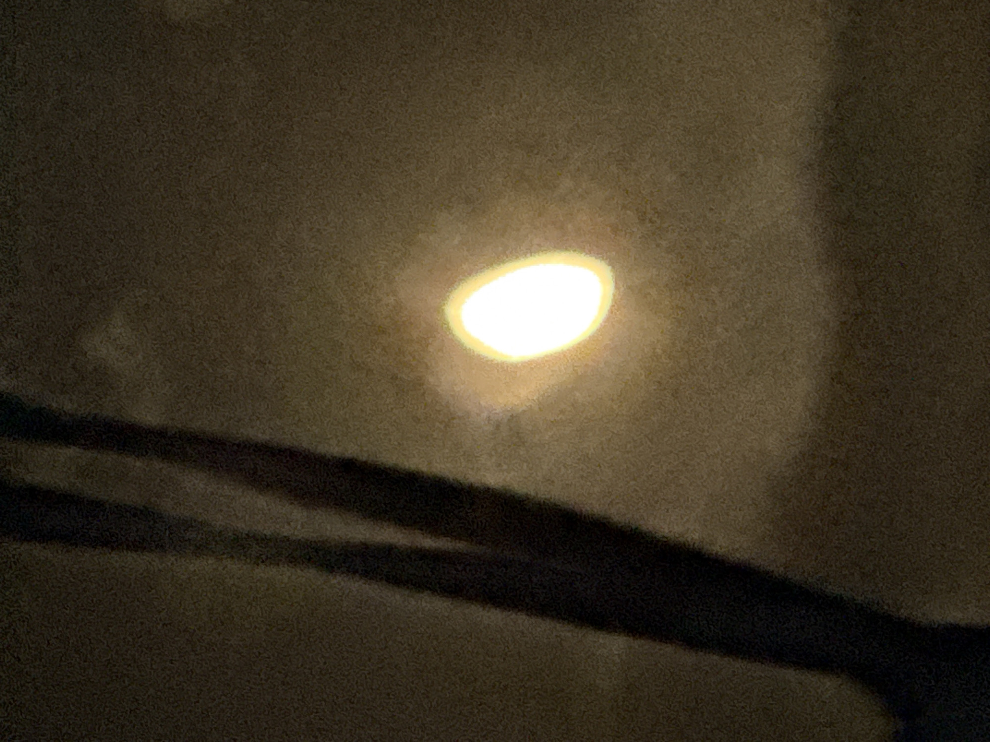 The image captures a dimly lit scene, likely at night, with a soft, diffused light source emanating from the center-right of the frame. This light source is oval-shaped and appears bright white with a yellowish halo, suggesting it could be a street lamp or a similar artificial light partially obscured by atmospheric conditions or a filter. To the left of the light source, two dark, vertical, elongated shapes cast shadows, implying the presence of objects such as poles, trees, or even structures. These shapes are largely indistinct due to the low lighting. The background is a uniform, textured dark brown, with subtle variations in tone that suggest a wall, sky, or other ambient surface. There are no visible people or overt actions. The overall impression is one of subdued illumination and a sense of stillness. The scene does not contain any identifiable landmarks or clues specific to Vostok, Kyrgyzstan.