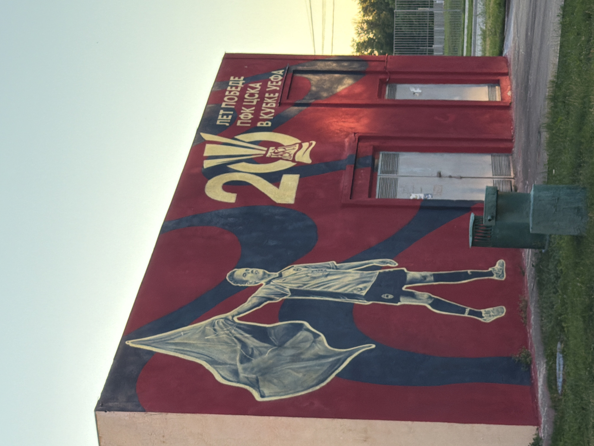 The image shows a mural on the side of a red building. The mural depicts a soccer player holding a flag, with the number "20" and Russian text in gold. The building has two metal doors and a window. In front of the building, there is a green trash can and a patch of grass. The sky is clear and light blue, and trees and a fence are visible in the background.